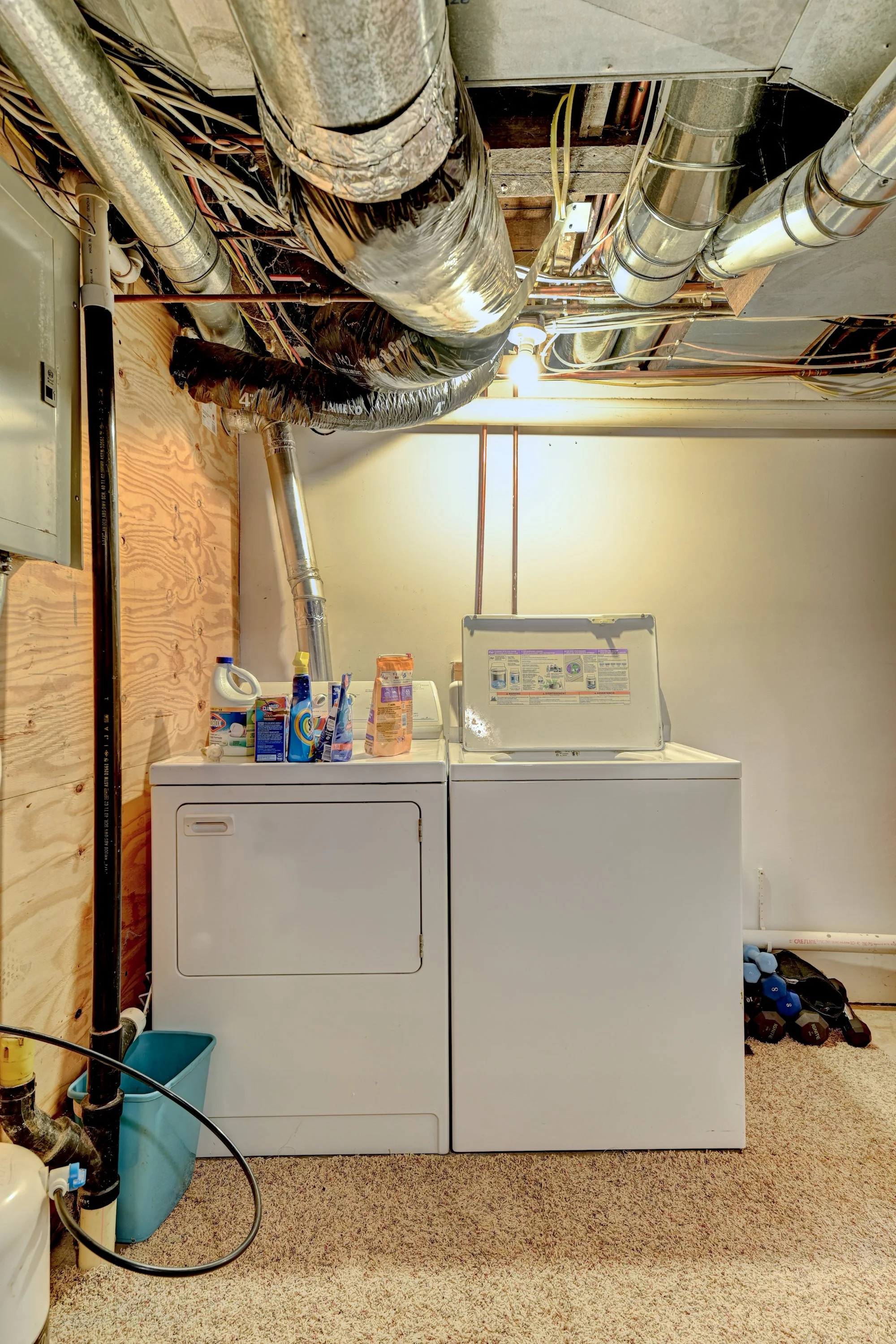 A laundry room with two white washing machines side by side, a wooden wall on the left with visible plumbing and electrical wiring, various laundry supplies on top of the machines, and a pile of dumbbells on the floor to the right.