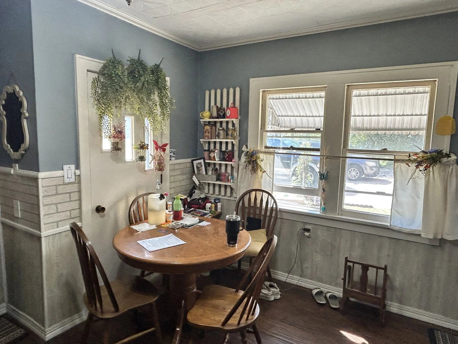 A kitchen or dining area with a round wooden table and four chairs. The table has papers, drinks, and various items on it. There are three windows with blinds and curtains, with some decorations hanging from the curtains. The walls are painted light 