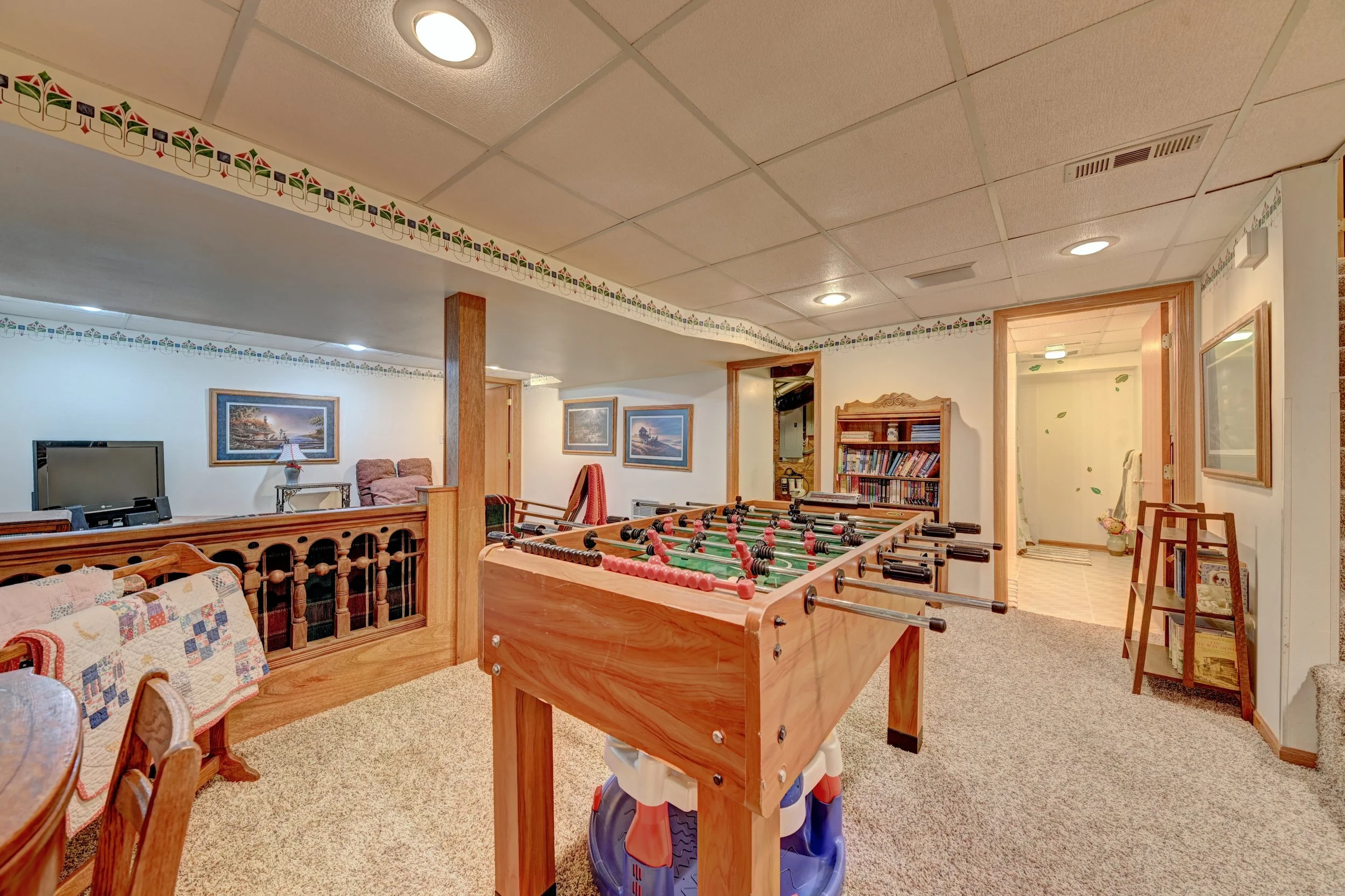 A cozy basement game room with a foosball table, a sofa with a quilt, and framed paintings on the wall, illuminated by ceiling lights.