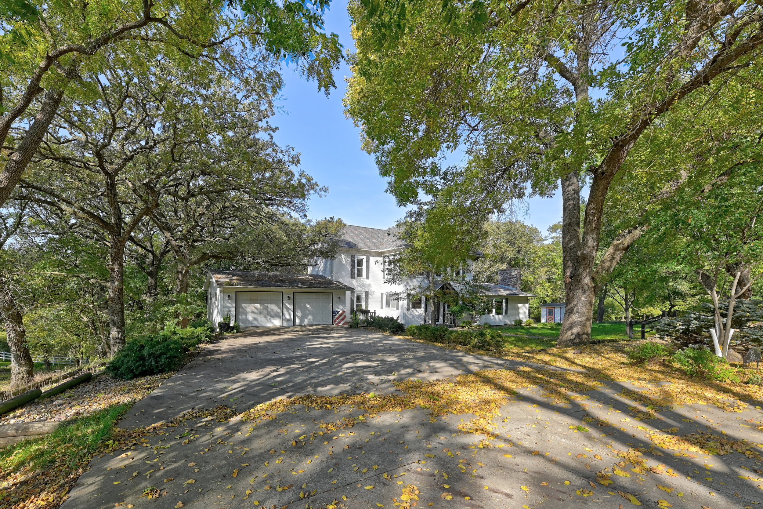 A driveway leading up to a white house with a garage, surrounded by tall trees with green leaves, some fallen yellow leaves on the ground, and a blue sky above.