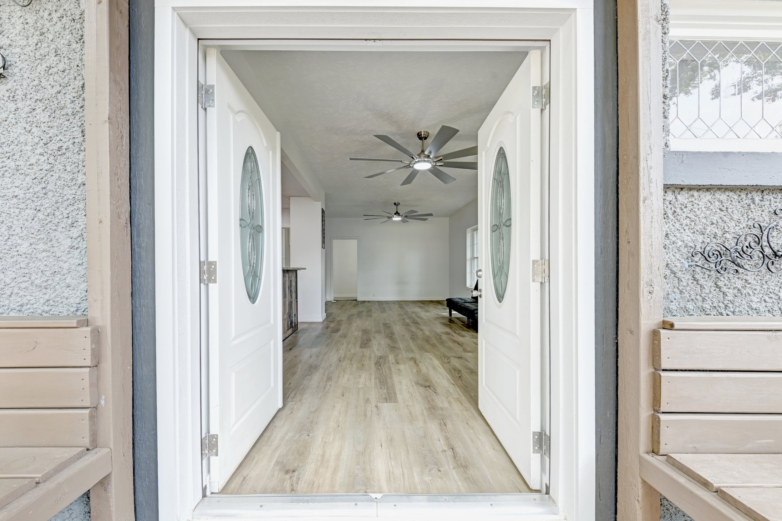 Open front door leading into a modern interior with light wood flooring, white walls, and ceiling fans, viewed from outside.