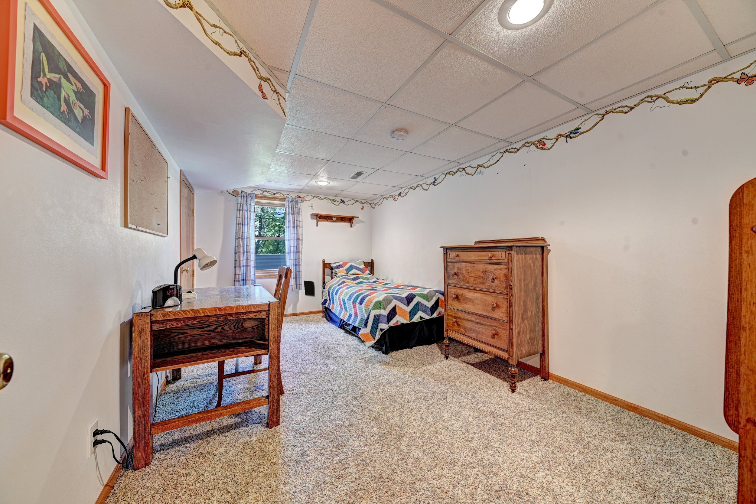 A bedroom with a bed covered by a colorful quilt, a wooden dresser, a window with plaid curtains, and wall decorations including framed pictures and a decorative branch running along the ceiling.