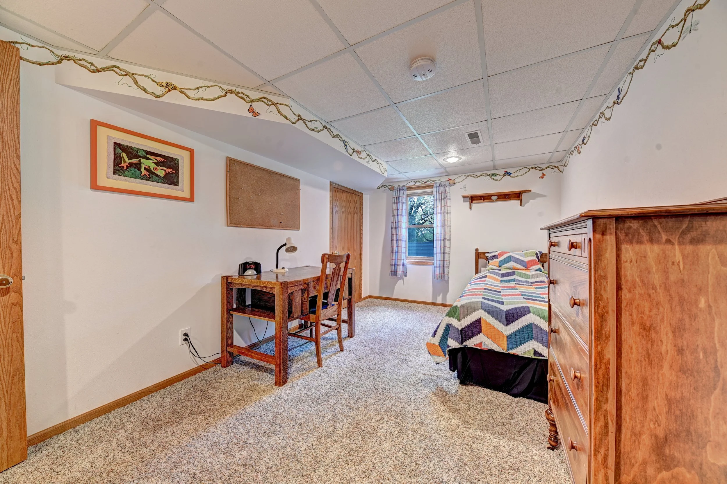 A bedroom with a bed covered in a colorful quilt, wooden furniture including a dresser, a small desk with a lamp, a chair, a window with plaid curtains, and decorations like a framed lizard picture and a vine with butterflies and insects on the ceili