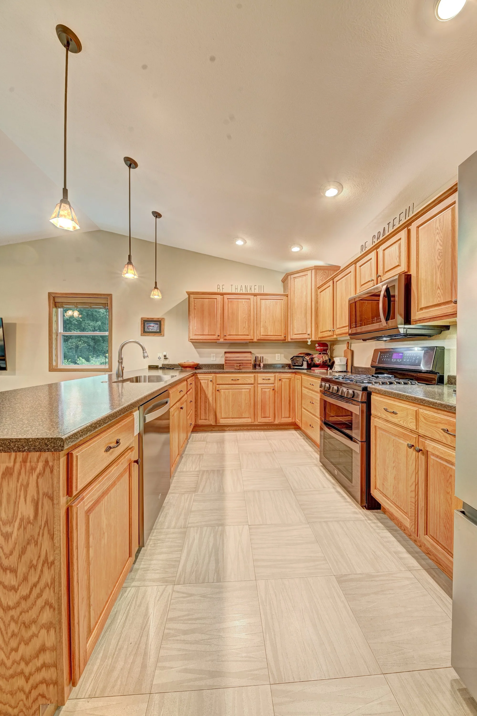 Kitchen with wooden cabinets, granite countertops, stainless steel appliances, pendant and recessed lighting, tiled floor, and a window showing greenery outside.
