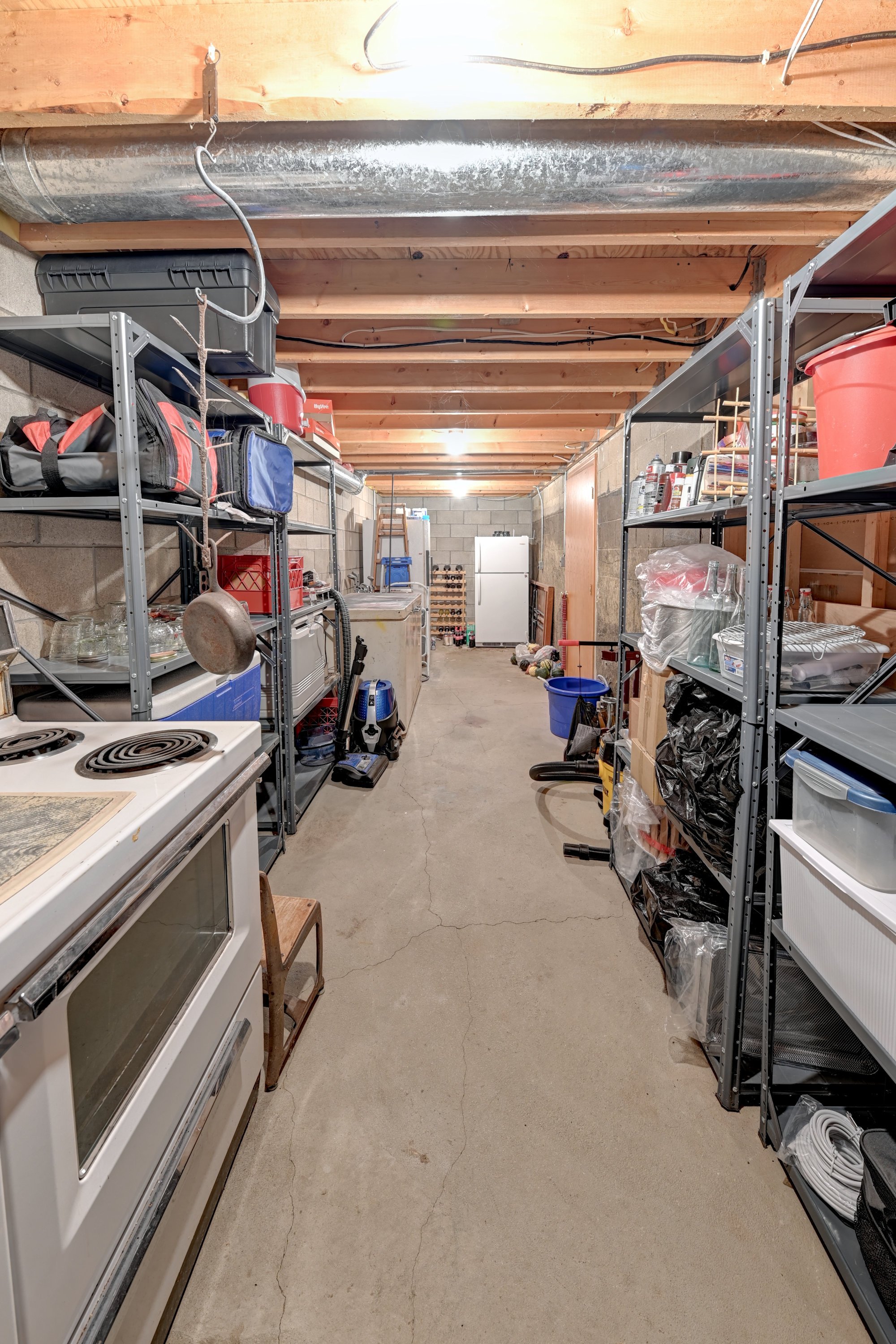 Inner view of a garage with metal shelving units holding various tools, supplies, and household items, concrete floor with visible cracks, unfinished wood ceiling, and a refrigerator at the back.