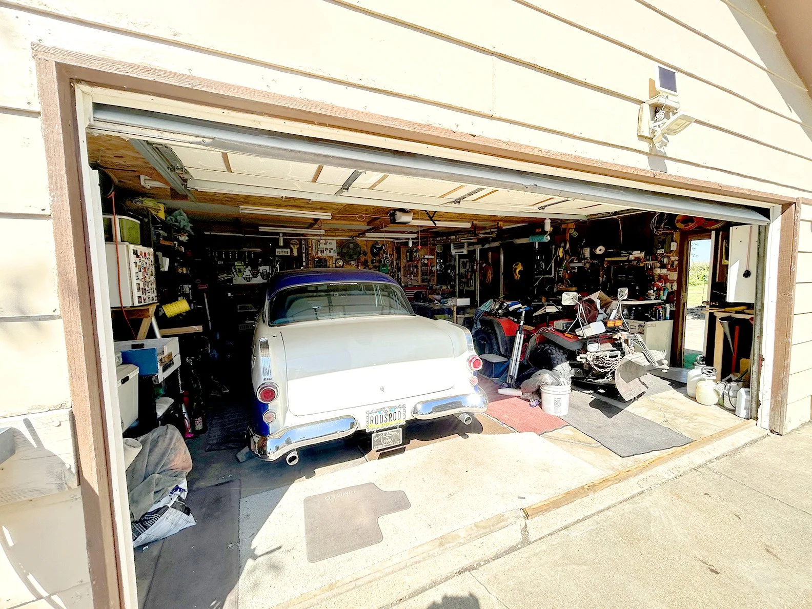 A garage with a classic car parked inside, along with various tools, equipment, and a motorcycle, with the garage door open.