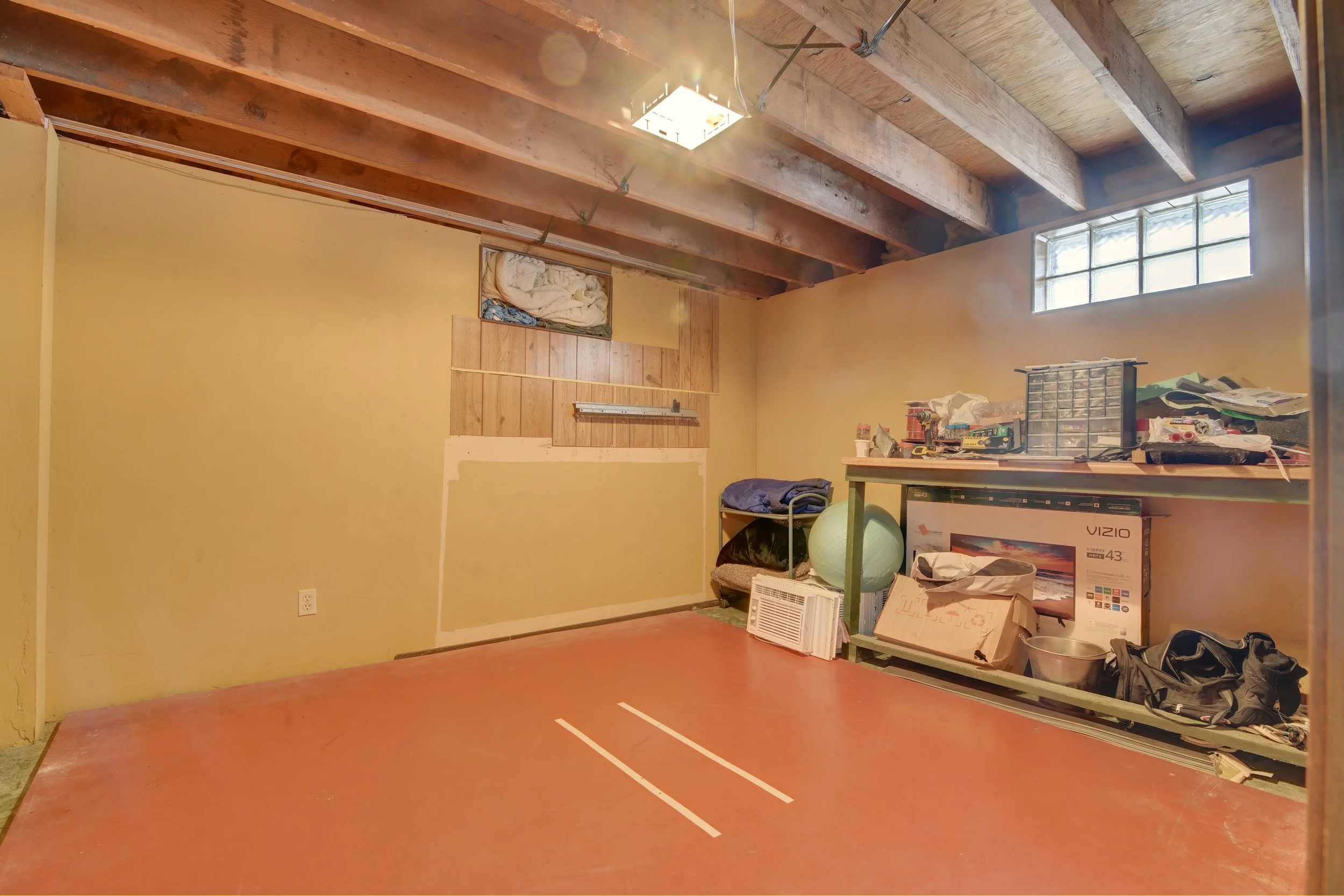 Empty garage or storage room with a concrete floor marked with two white lines, unfinished walls, and an exposed wooden beam ceiling. There is a small window near the ceiling and a shelf with various items including boxes, cleaning supplies, and an e
