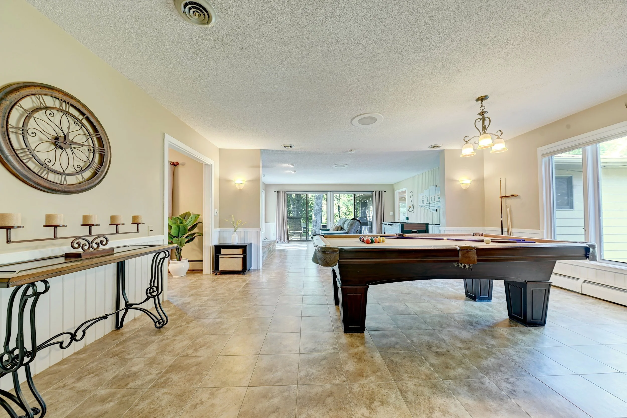 A spacious living room with tile flooring, a pool table, a large wall clock, a chandelier, and large windows leading to a screened porch.