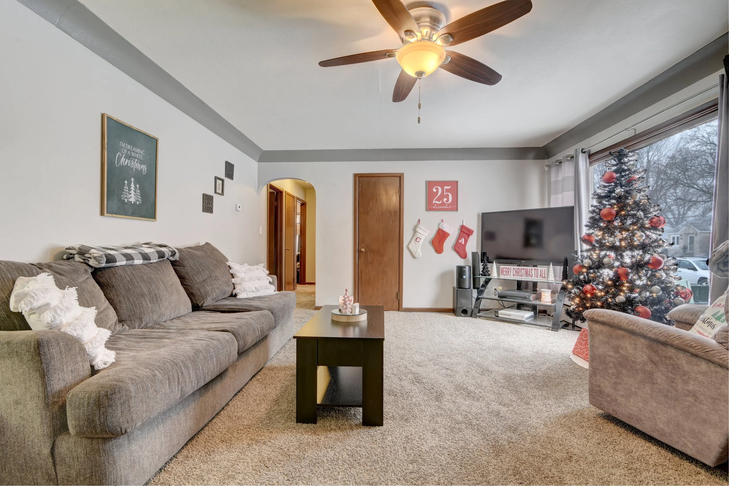 Living room decorated for Christmas with a tree adorned with red ornaments and lights, a couch with pillows, a TV on a stand, a coffee table, and holiday decorations on the walls.