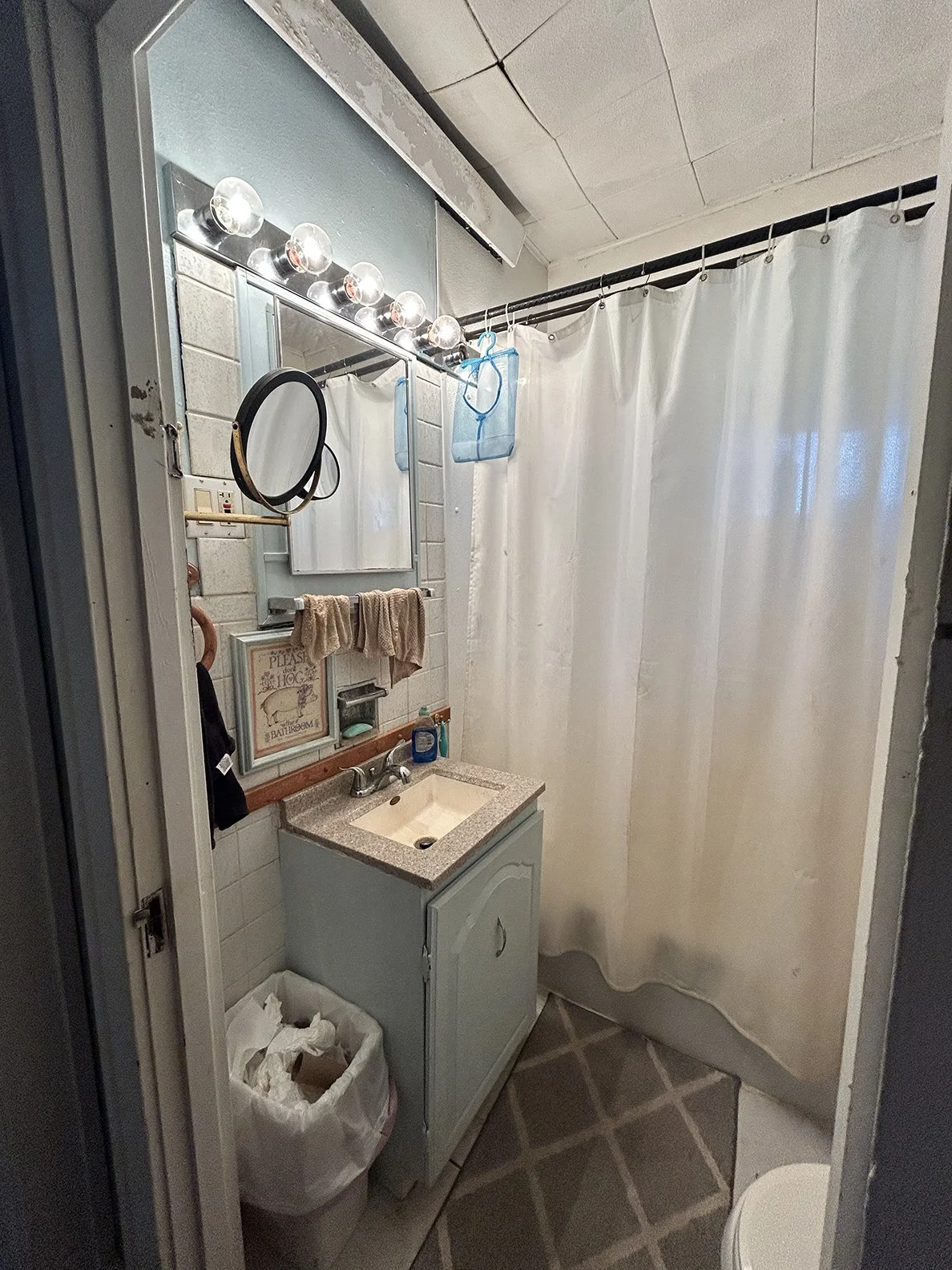 A small bathroom with a vanity mirror, a countertop sink, and a shower with a white curtain. There are four round bulbs above the mirror, a black hand mirror, and a sign on the wall that says "Please don't hog the bathroom." A blue mesh laundry bag h