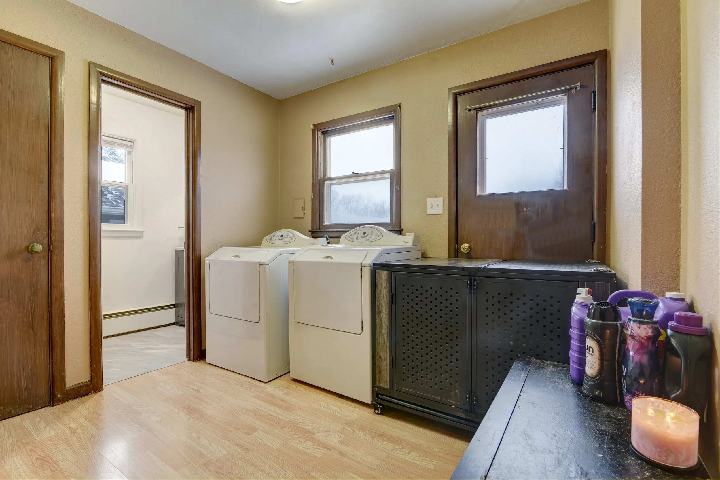 A laundry room with beige walls, wooden trim, a washer and dryer, a black storage cabinet, some detergent bottles, and a candle on a black surface.