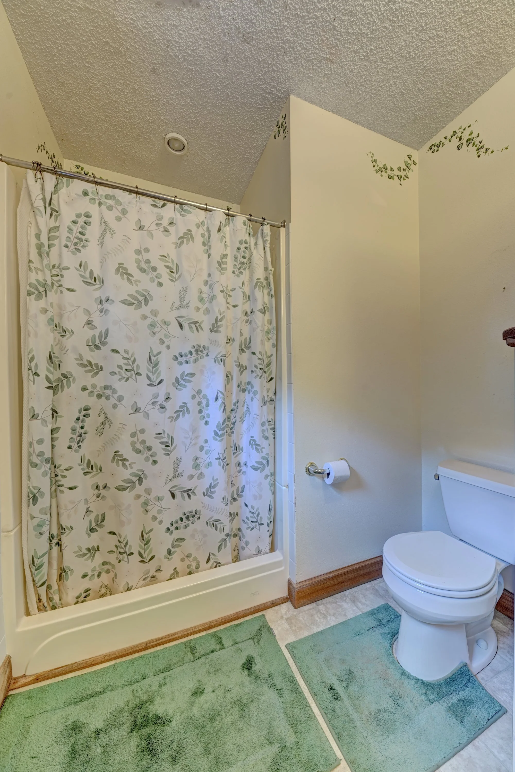 A bathroom with a shower area covered by a white curtain with green leaf patterns, a toilet, a green rug on the light-colored tile floor, and small decorative green leaf designs on the walls.