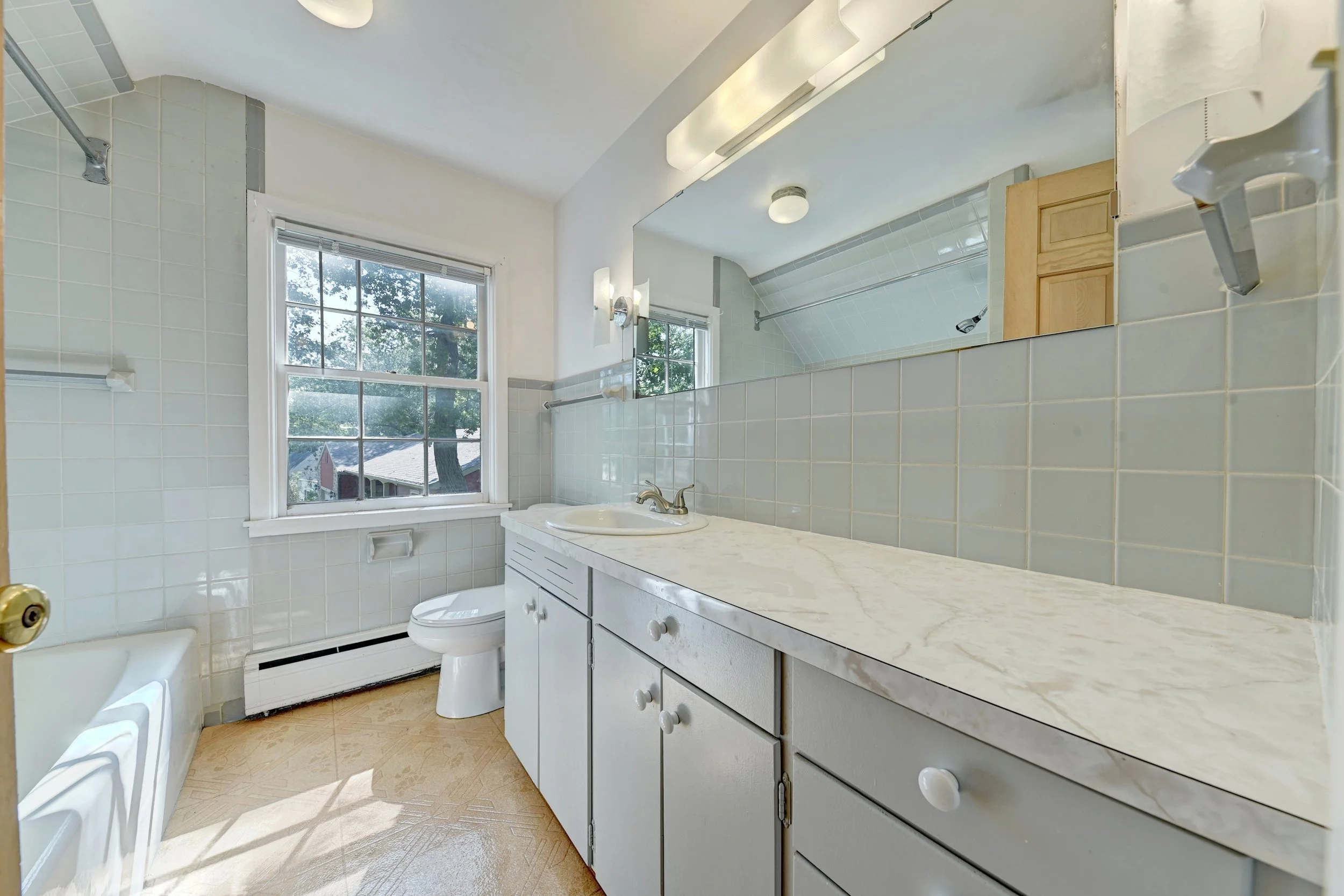 A bathroom with a large window, white tiles on the walls, a marble countertop with a sink, a mirror reflecting the room, a bathtub partially visible, and natural light streaming in.