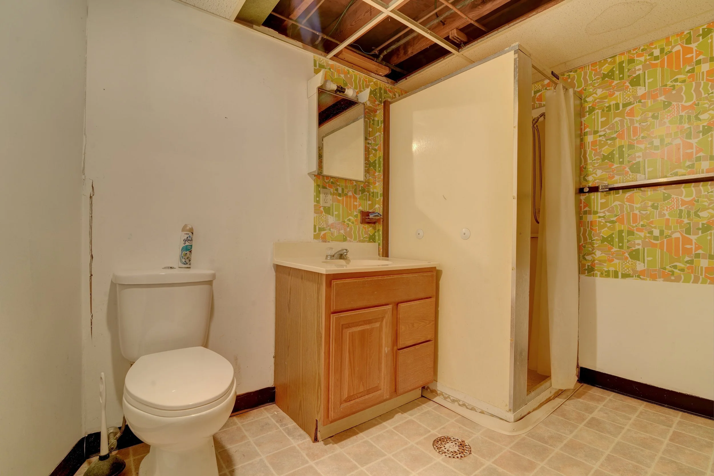 Bathroom with a toilet, a small wooden vanity with a mirror above it, and a shower stall with a curtain. The walls have colorful, patterned wallpaper, and the ceiling is partially open, exposing ceiling tiles and wiring.