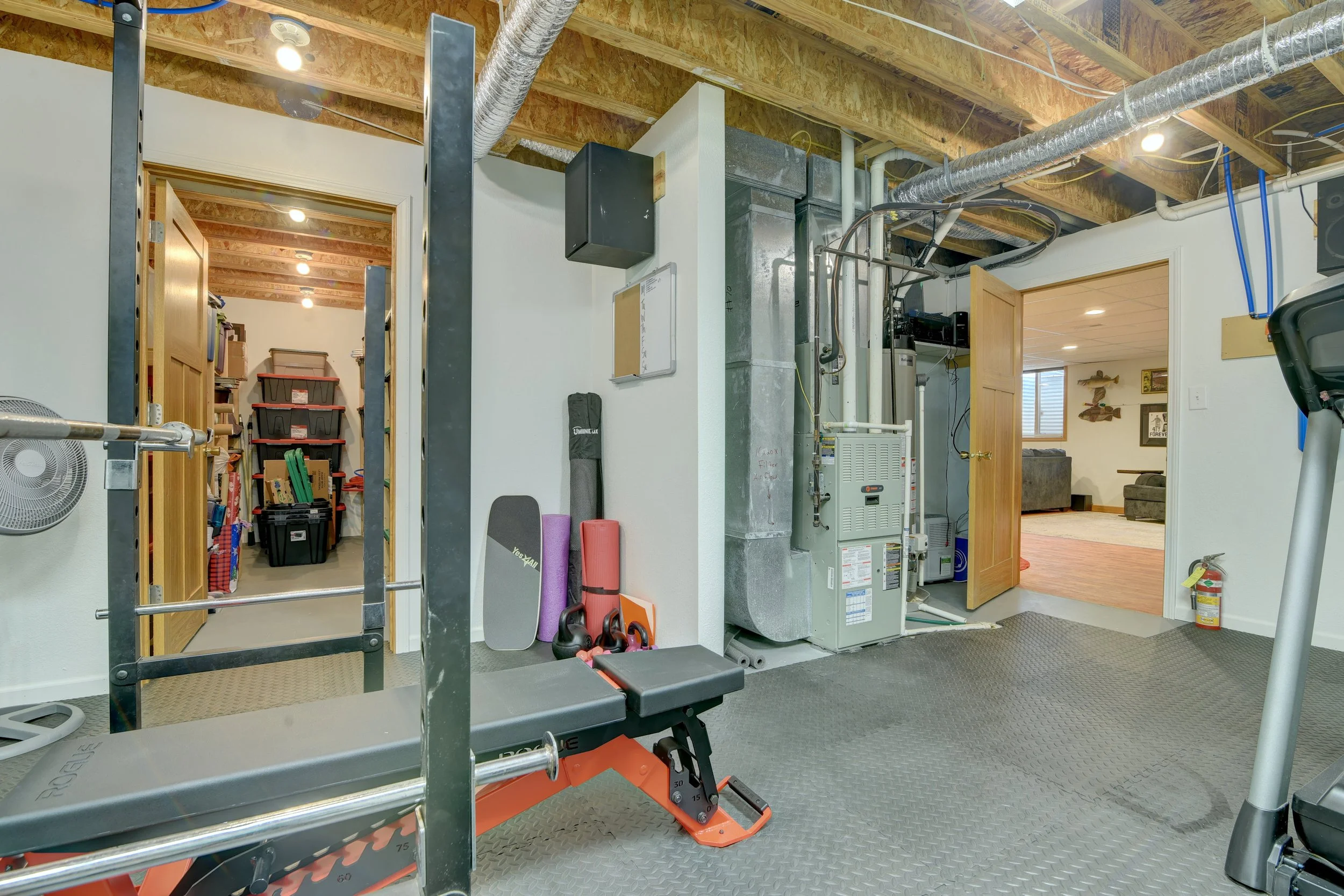 Home gym and storage room with exercise equipment, kettlebells, mats, storage shelves, and utility heater, adjacent to living room.