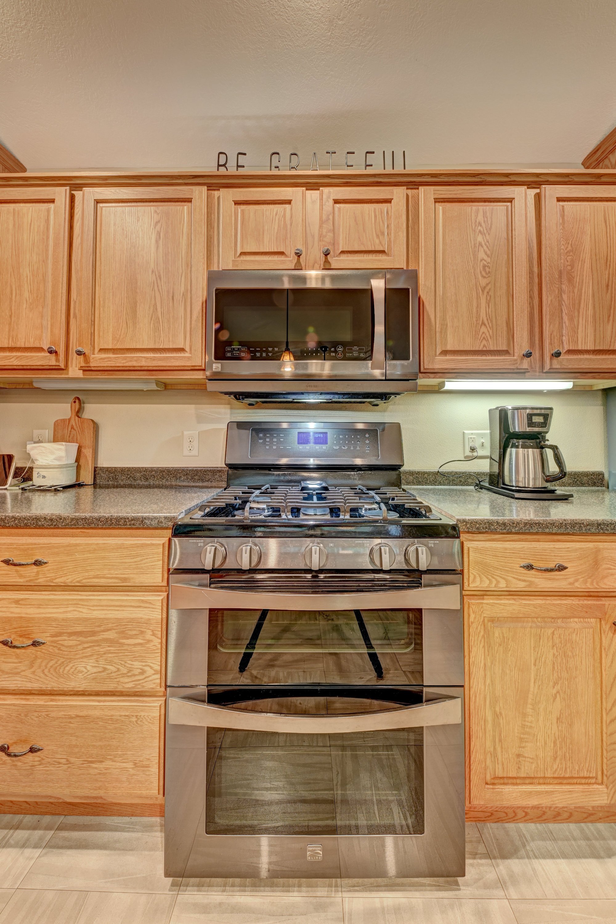 Kitchen with wooden cabinets, stainless steel microwave and stove, coffee maker, tissue box, cutting board, and a sign above saying 'BE GRATEFUL'.