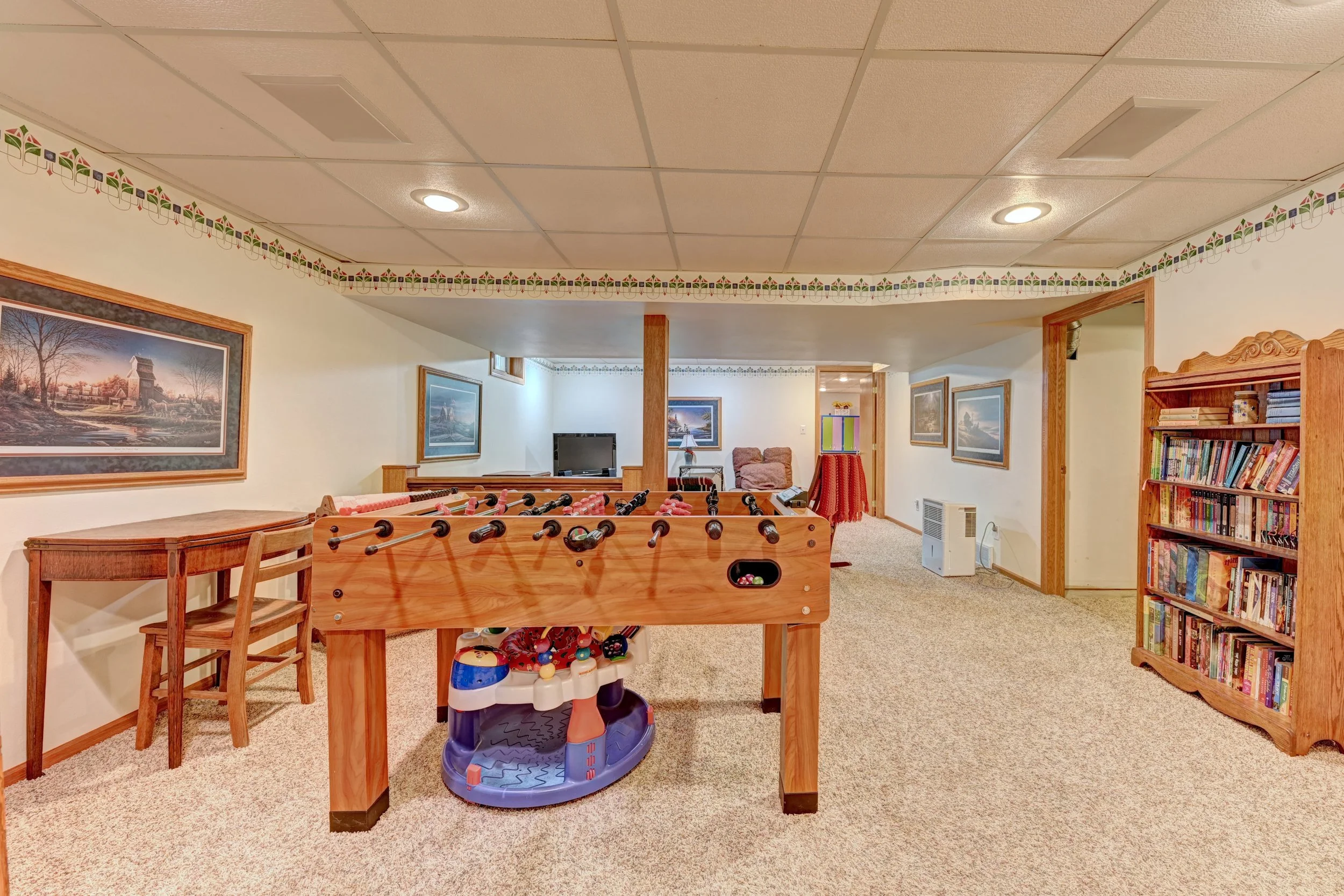 A cozy basement recreation room with a foosball table, bookshelf, armchair, tv, and framed landscape paintings, decorated with a patterned border near the ceiling.