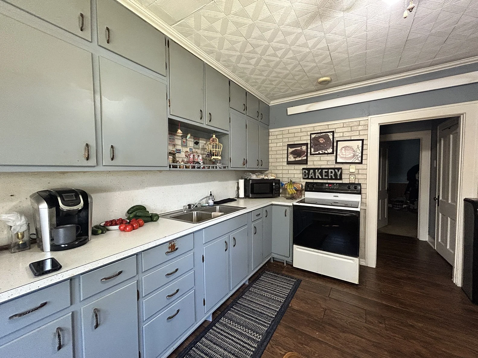 A kitchen with blue cabinets, white countertops, a stainless steel coffee maker, fresh vegetables on the counter, a microwave, a stove with an oven, and a wall sign that says 'BAKERY' above the stove. There are three framed pictures on the wall, and 