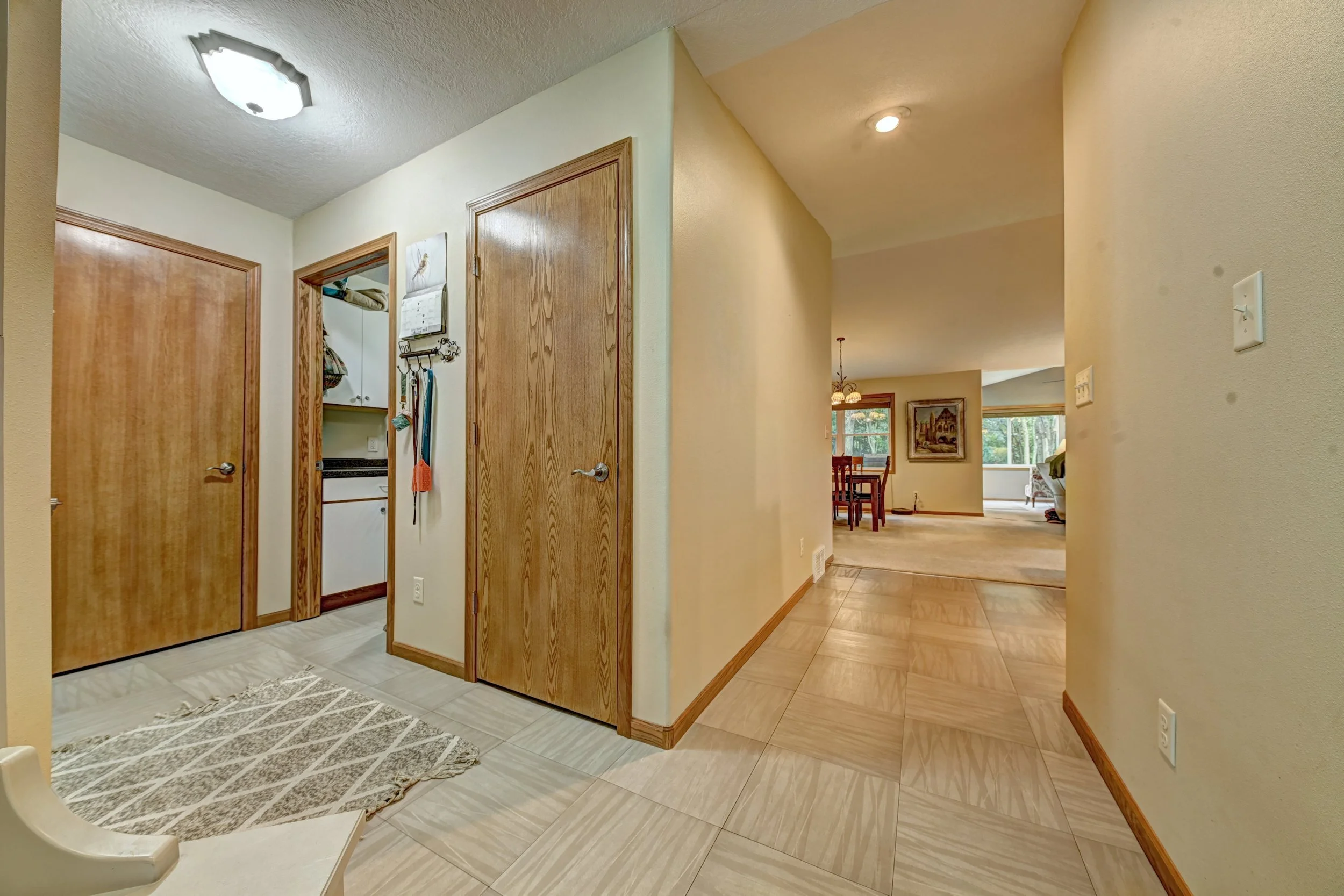 Interior view of a home featuring a hallway with a wooden front door, a closet with wooden doors, and a kitchen area visible through an open doorway. The space has beige walls, wooden trim, and wood-look flooring, with natural light coming from the w