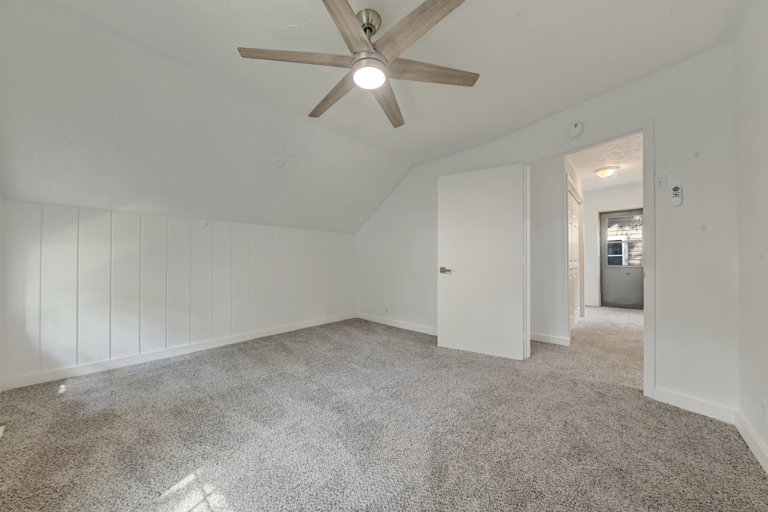 Empty room with white walls and beige carpet, ceiling fan with light, open door to hallway, window in hallway.