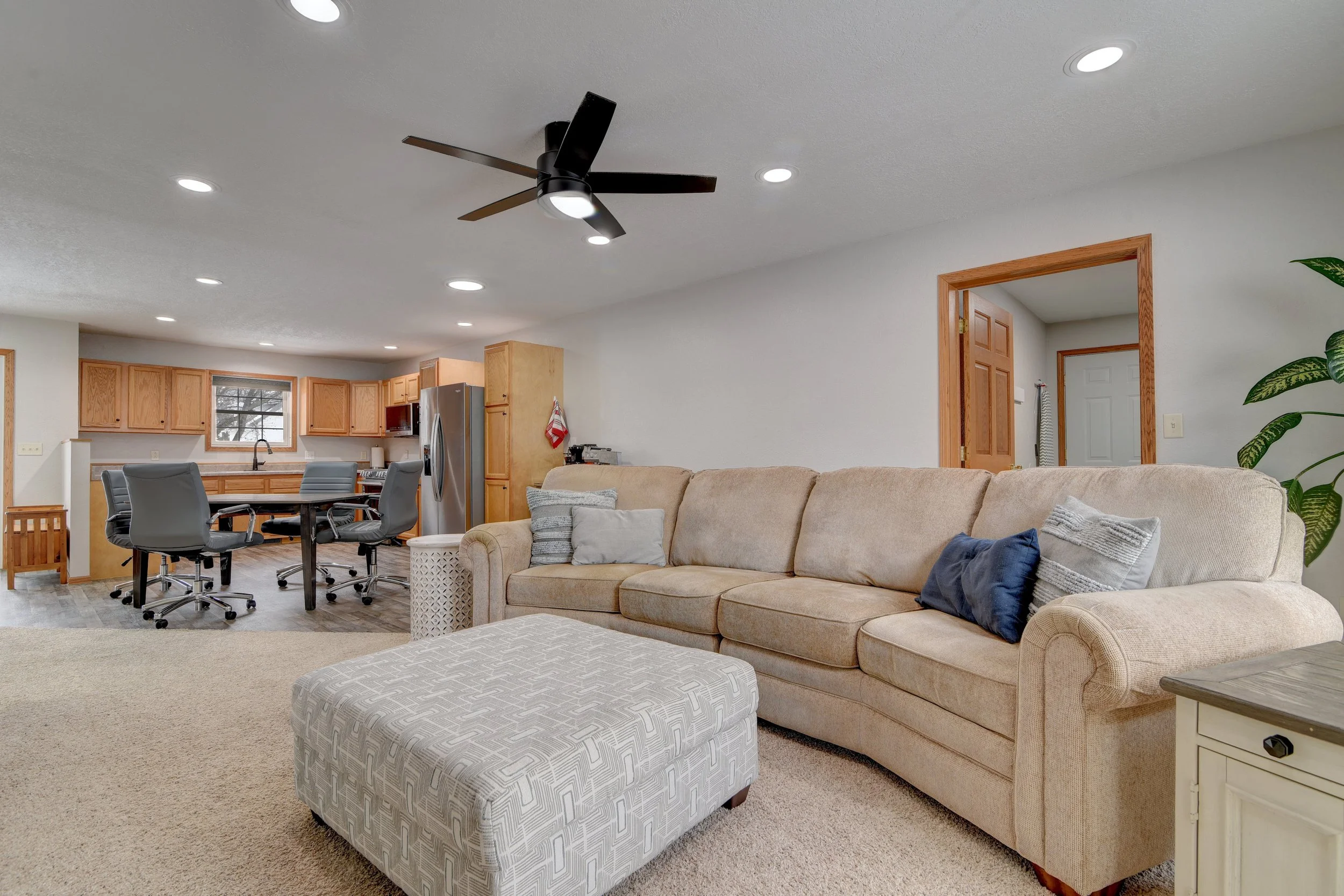 Living room and kitchen area with a beige sectional sofa, ottoman, several pillows, a ceiling fan, and a dining table with chairs in an open floor plan.