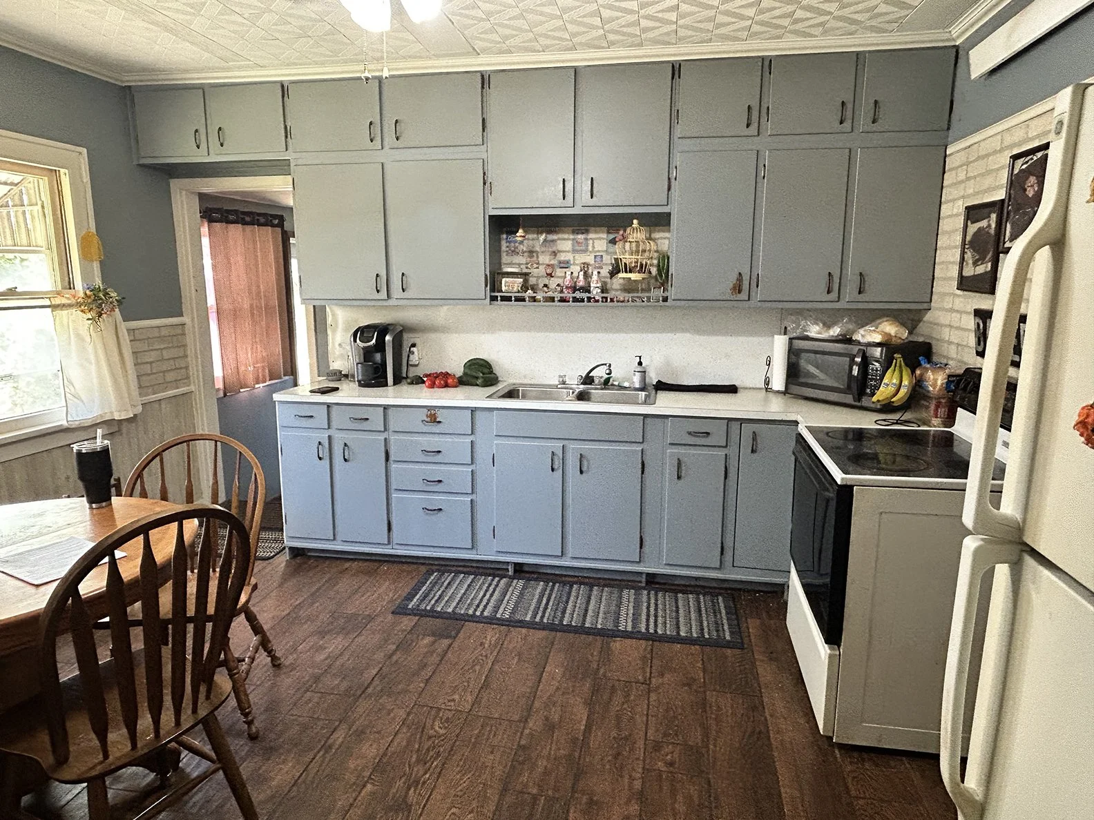 Kitchen with blue cabinets, wooden dining table with chairs, window with curtains, white and black stove, microwave, coffee maker, and fresh produce on the counter, hardwood floor, and wall decor.