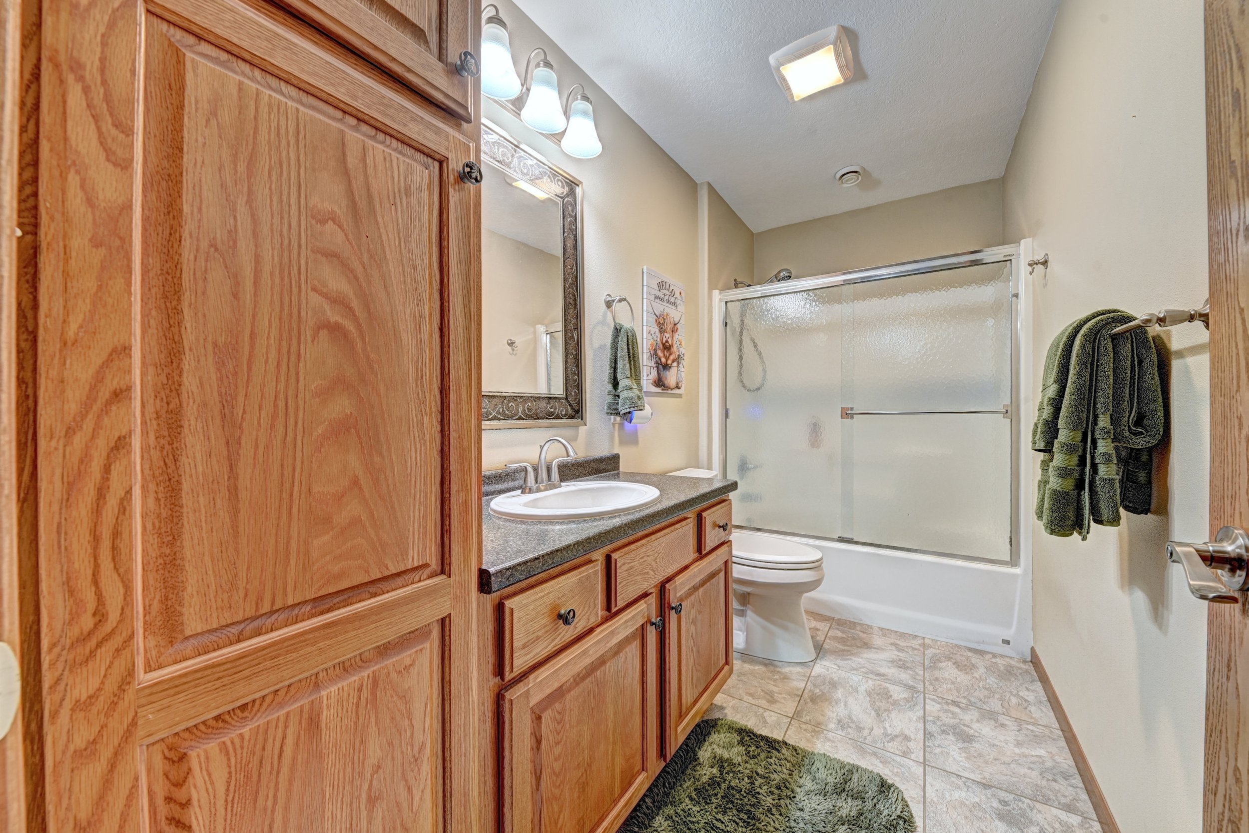 A small bathroom with wooden cabinets, a mirror, a sink with a granite countertop, a toilet, and a shower with a frosted glass sliding door. The walls are painted in a light beige color. There are green towels hanging on a towel bar and a small mat o