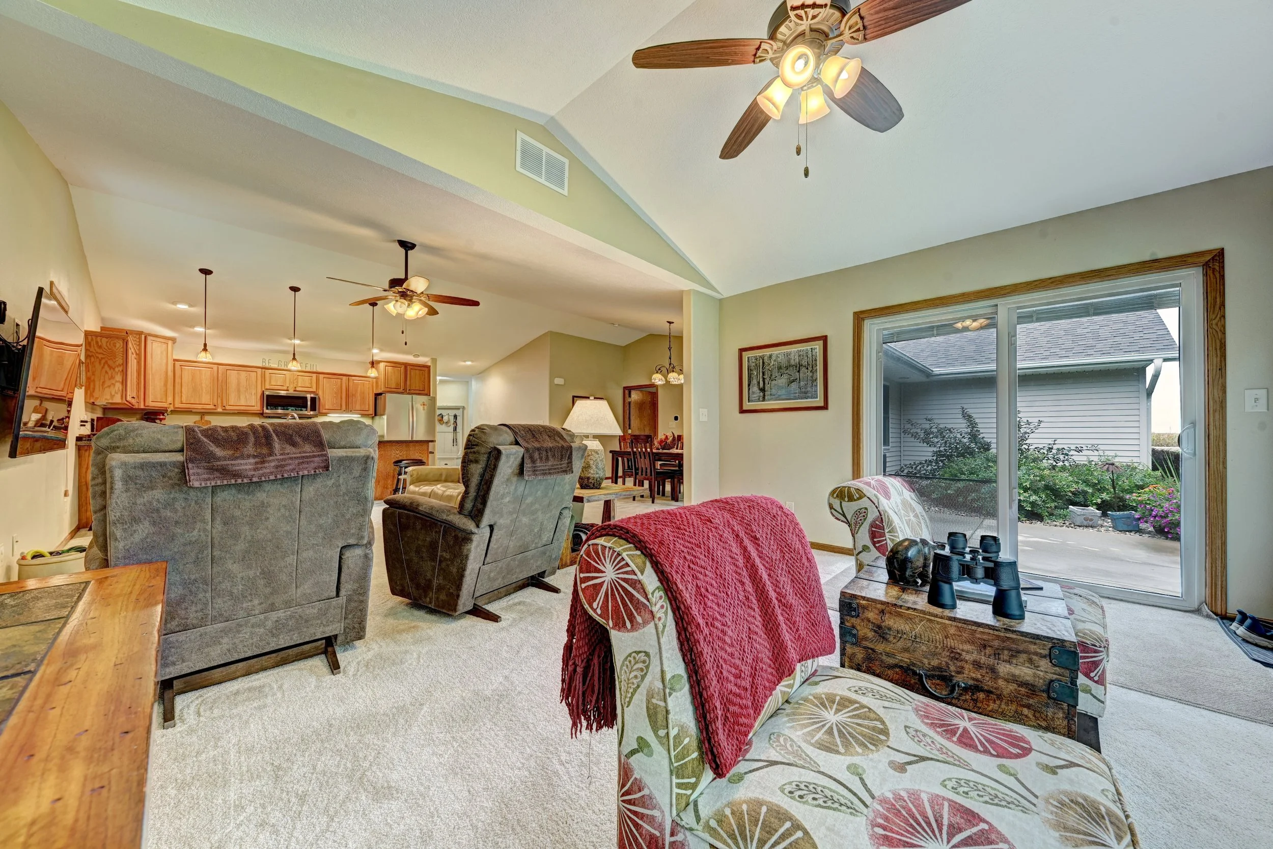 Living room area with beige carpet, multiple chairs, wooden coffee table, large sliding glass door leading outside, kitchen with wooden cabinets and pendant lighting, ceiling fans, and natural light.