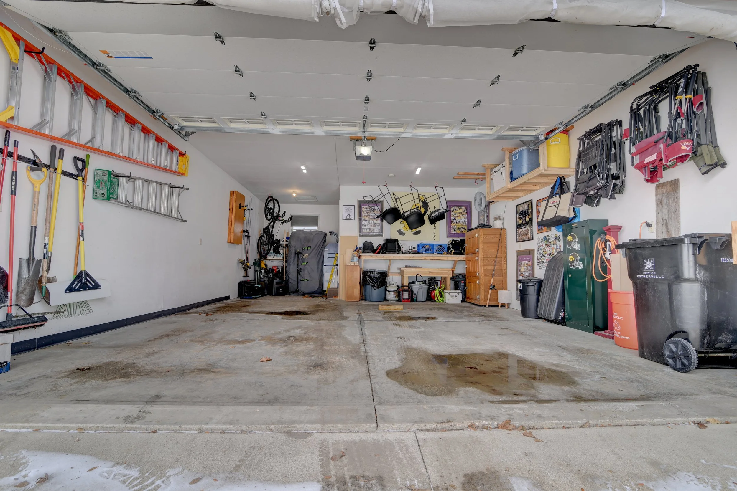 Empty garage with tools and storage on the walls, some wet spots on the concrete floor, and various stored items on shelves and in trash bins.