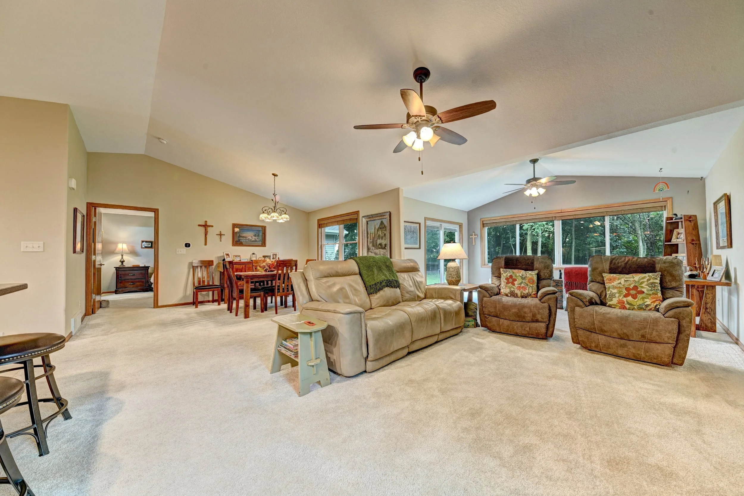 Spacious living room with beige carpet, multiple sofas, large windows, ceiling fans, wooden dining table, and decorative religious and artwork on walls.