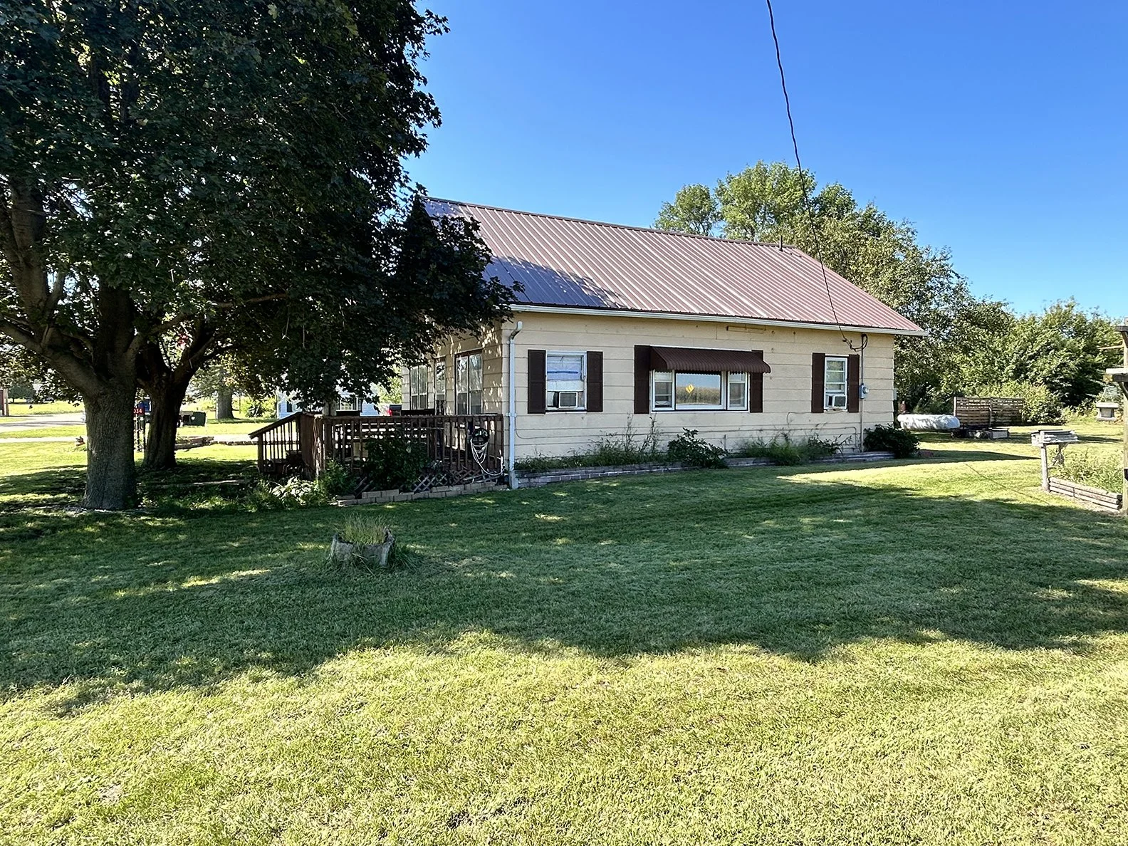 A single-story house with light-colored siding, dark shutters, and a metal roof, surrounded by green grass and trees on a sunny day.