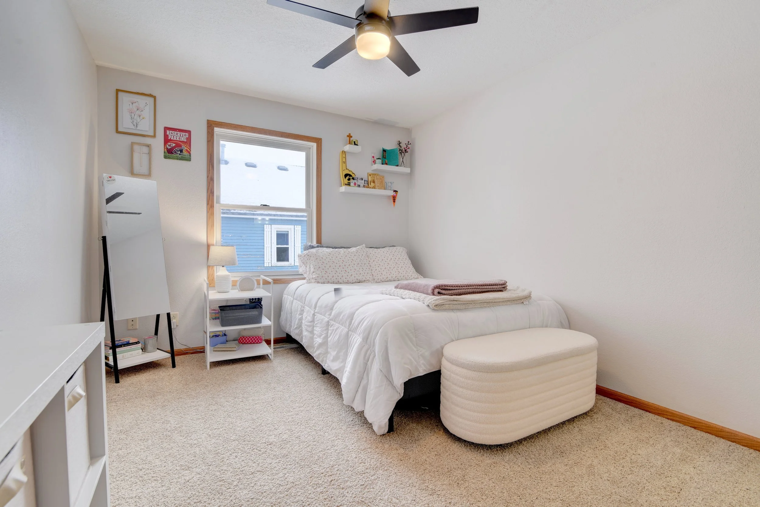 A bedroom with white walls, a window with a wooden frame, a white bed with pillows and folded blankets, a white ottoman at the foot of the bed, a white nightstand with a lamp, a full-length mirror, and floating shelves with decorations. There's a bei