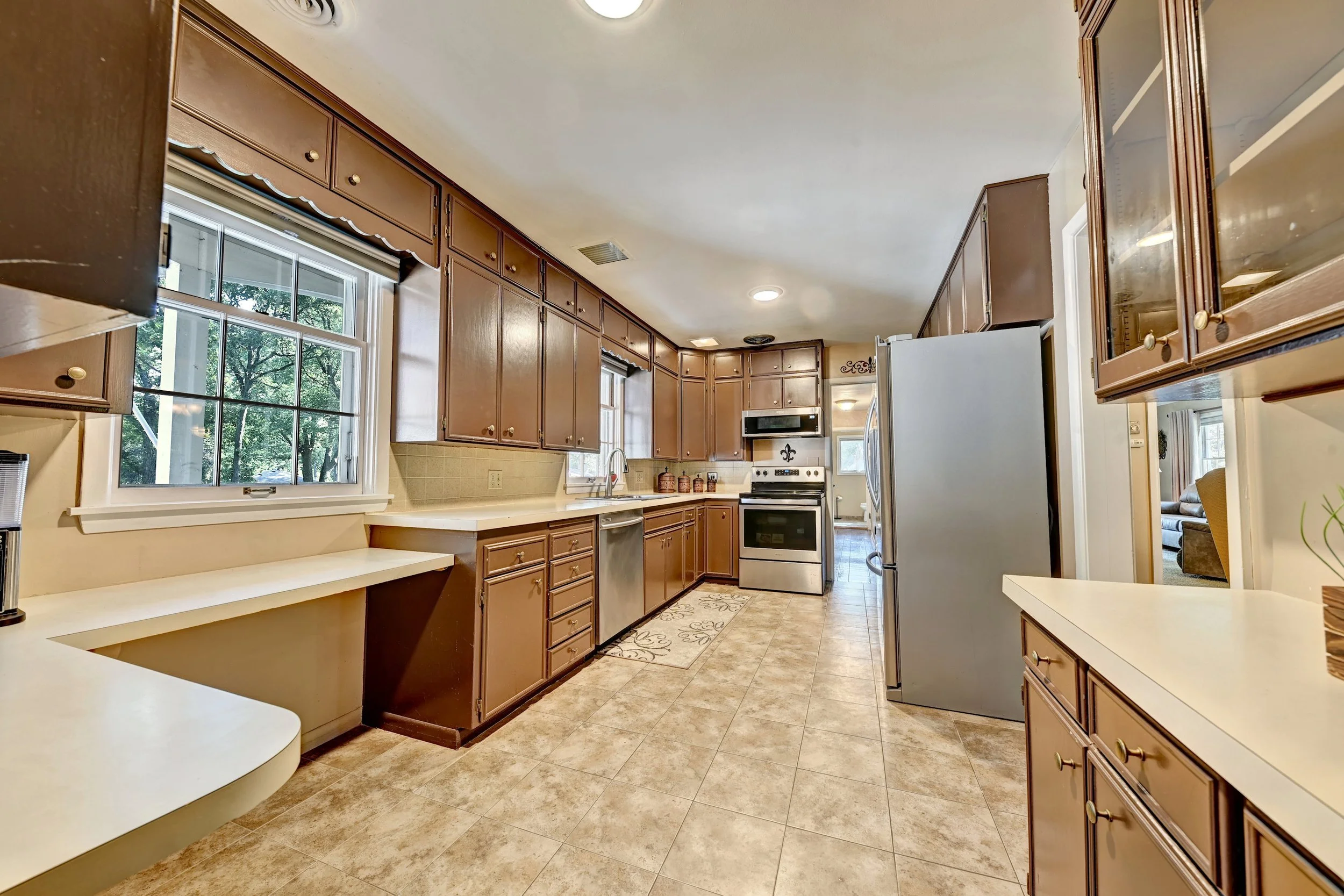 Kitchen with wooden cabinets, tile floor, and appliances including a stove, microwave, and refrigerator. Large window shows trees outside.