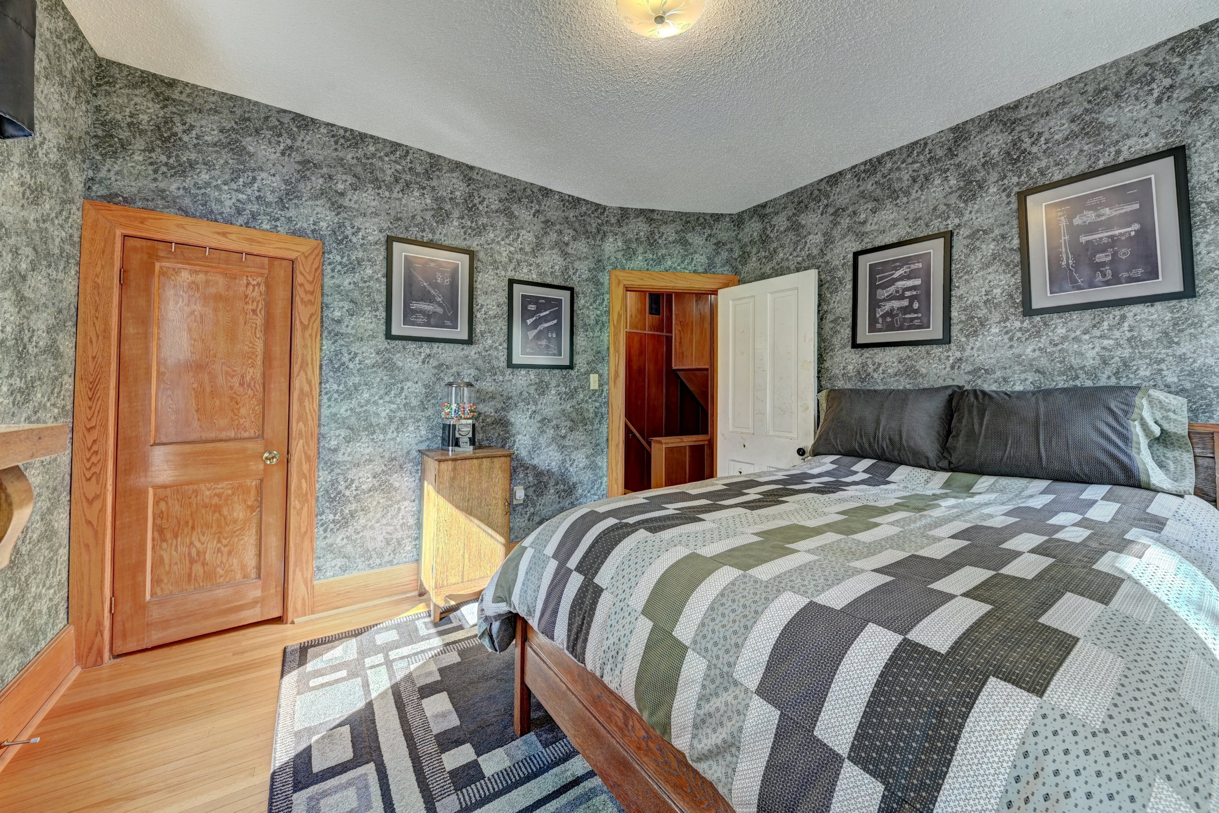 A bedroom with textured gray walls, a wooden door, a small nightstand, a bed with a patchwork quilt, and framed blueprint artwork on the walls. There is a gumball machine on the nightstand and sunlight coming in from the left.