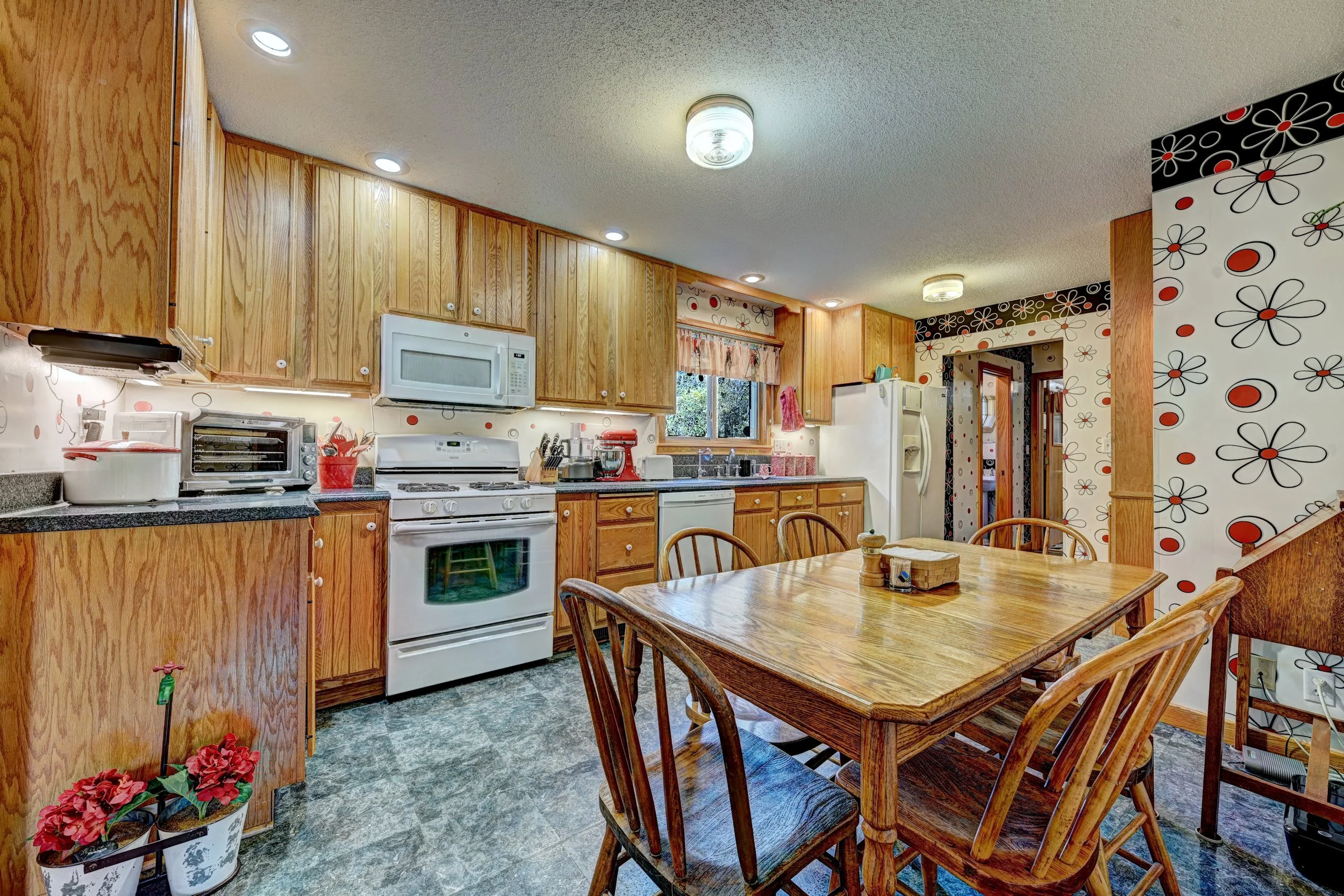 A kitchen with wooden cabinets, a white stove, microwave, and refrigerator. There is a round wooden dining table with wooden chairs and decorative wallpaper with black, white, and red flowers.