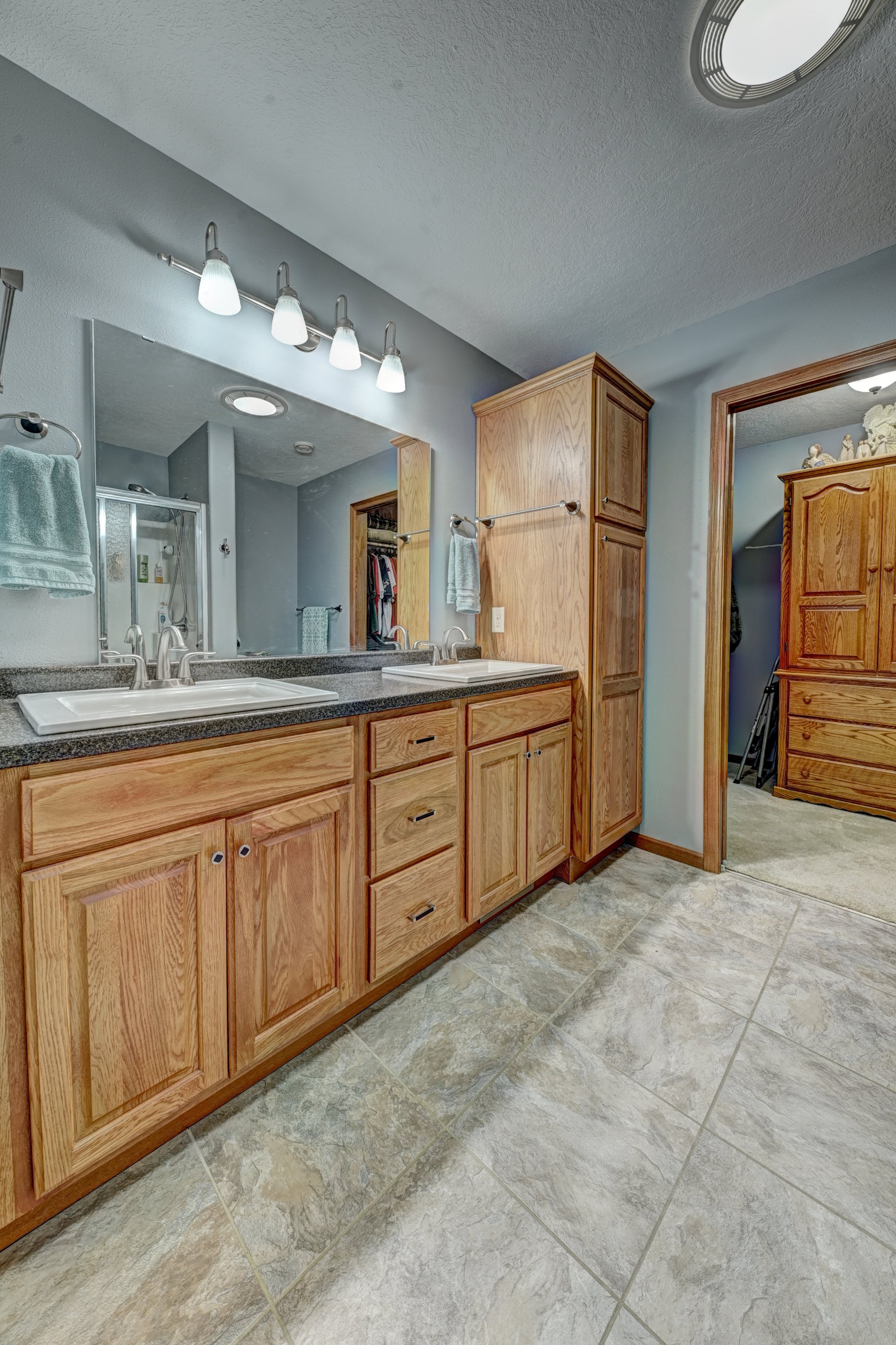 A bathroom with wooden cabinets, a double sink vanity, a large mirror, and tiled floor. There are light fixtures above the mirror and various bathroom items visible in the reflection.