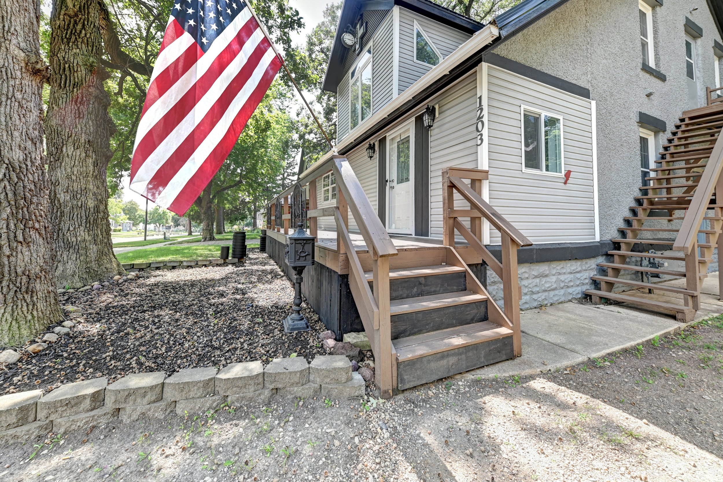 Front porch of a house with wooden stairs, a white door, black lantern lights, a large American flag hanging from a tree, and mature trees in the background.