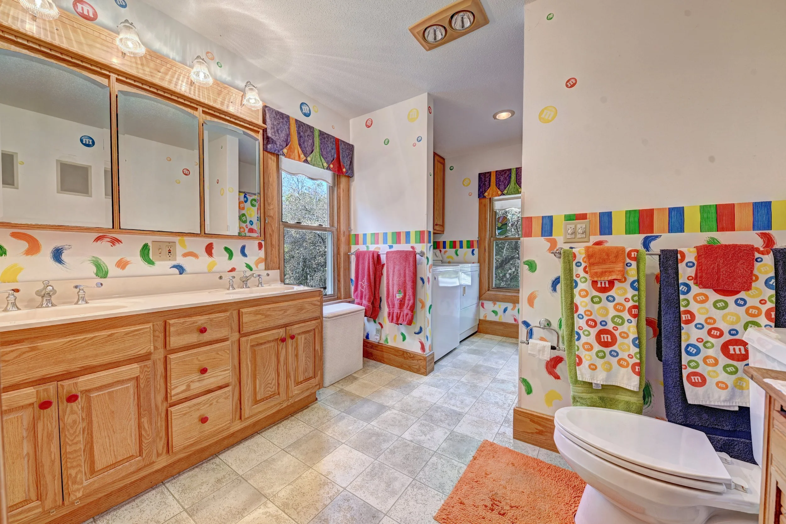 A colorful bathroom decorated with M&M's themed stickers and curtains, featuring a wood vanity with a large mirror, a window with a scenic view, and towels hanging near a washing machine.