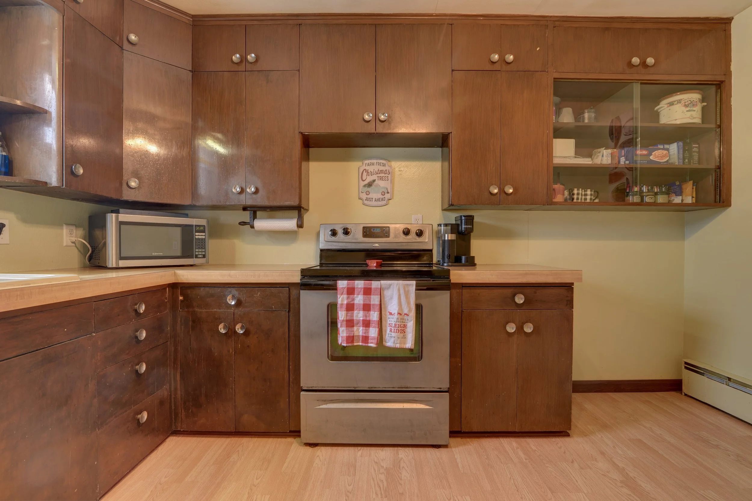 A kitchen with wooden cabinets, a stainless steel oven with two dish towels hanging on its door, a microwave oven on the counter, and a coffee maker next to the oven. There is a sign on the wall that says "Farm Fresh Christmas Trees Just Ahead." A gl