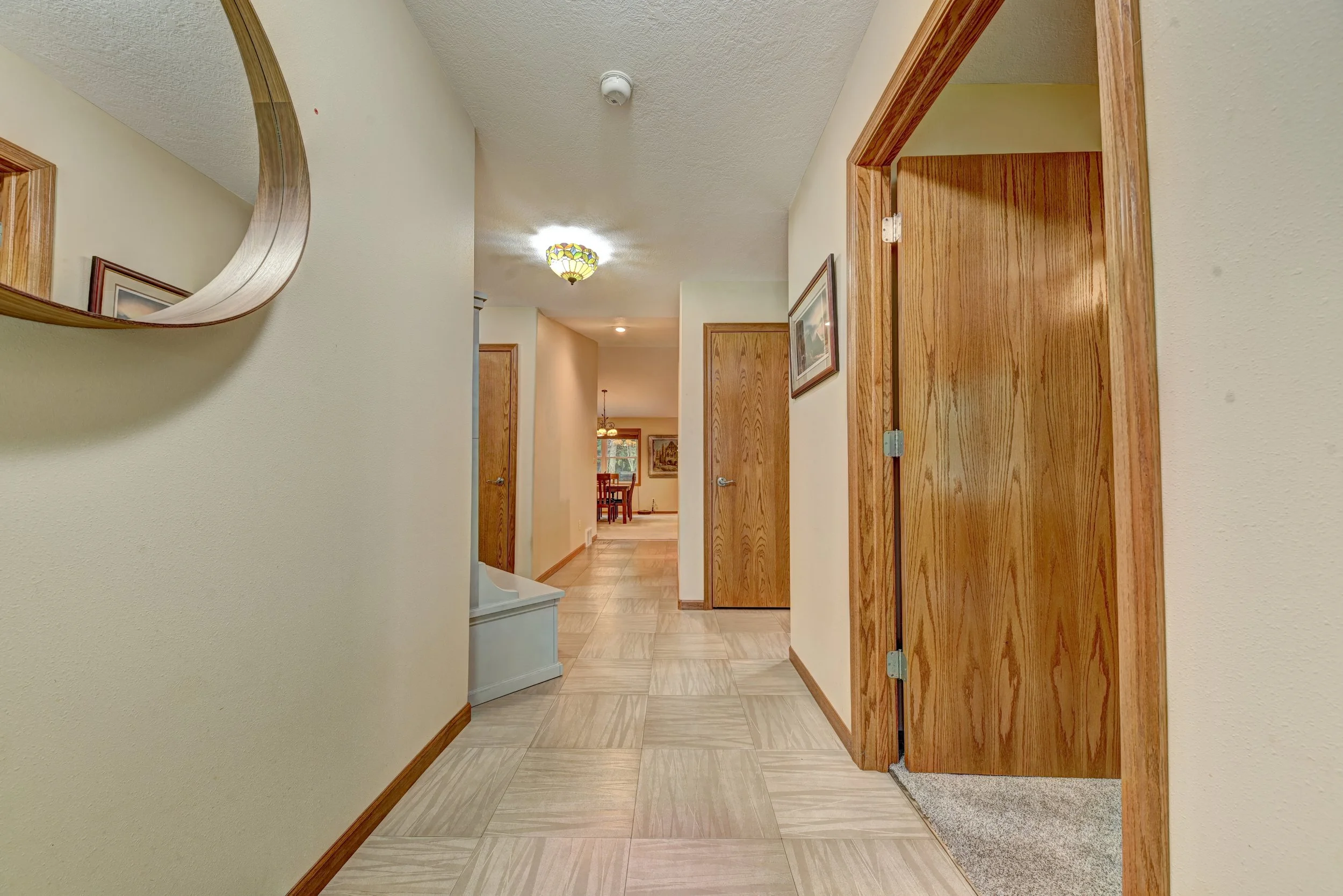 View of a hallway in a home with light-colored walls, wooden trim, and tiled flooring. Part of a rounded mirror is visible on the left wall, and an open doorway with an brown wooden door frame is on the right. In the background, a dining area with a 