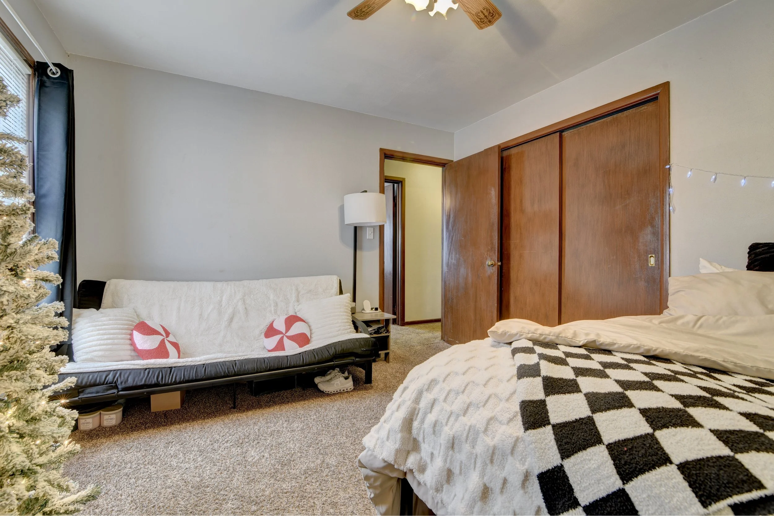 A bedroom with a bed covered in a black-and-white checkered blanket, a black futon with holiday-themed pillows, a side table with a lamp, a lamp on the wall, and a Christmas tree partially visible on the left. The room has beige carpet, a ceiling fan