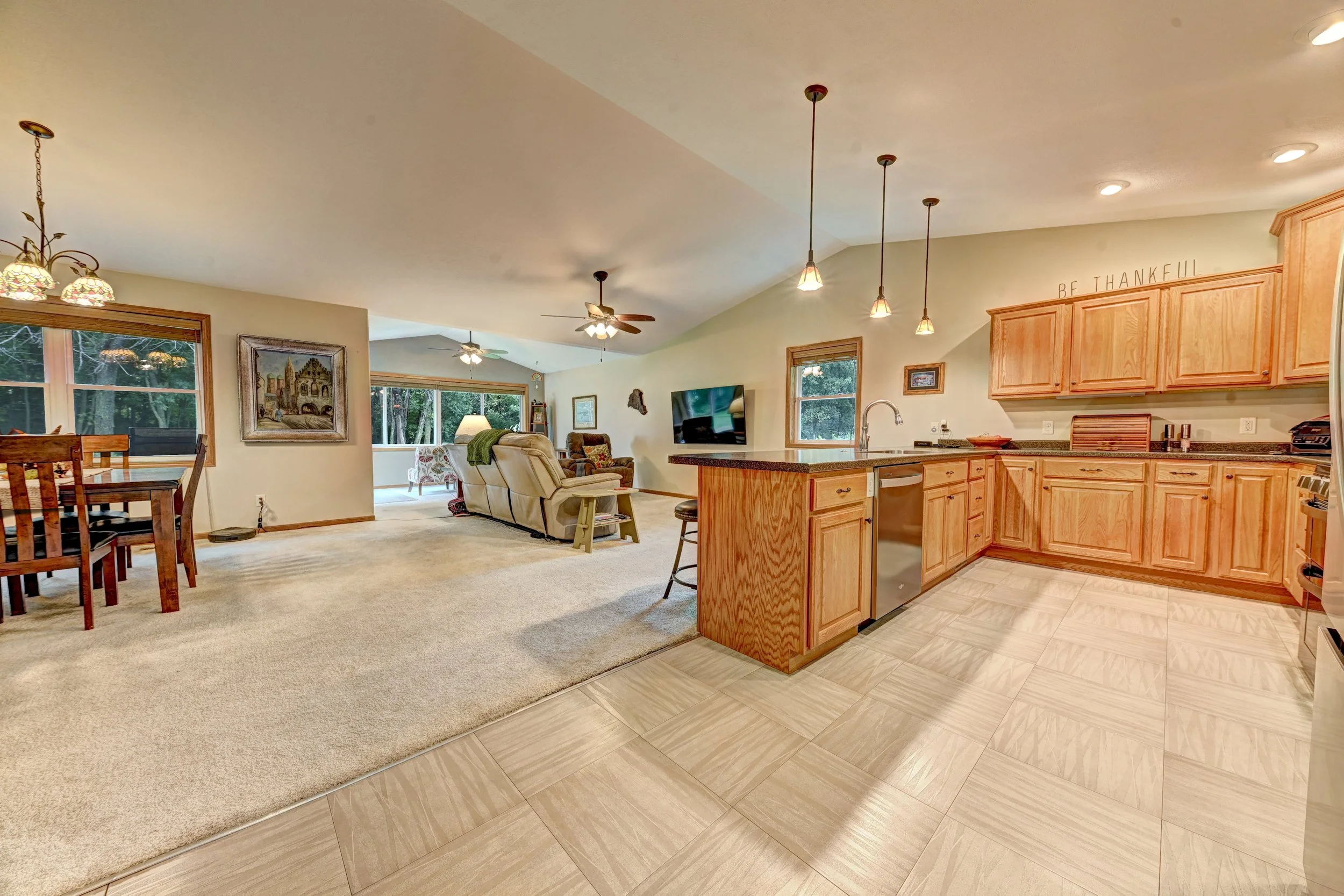 Open-concept living room and kitchen with wood cabinets, ceiling fans, pendant lights, and a dining area with large windows.