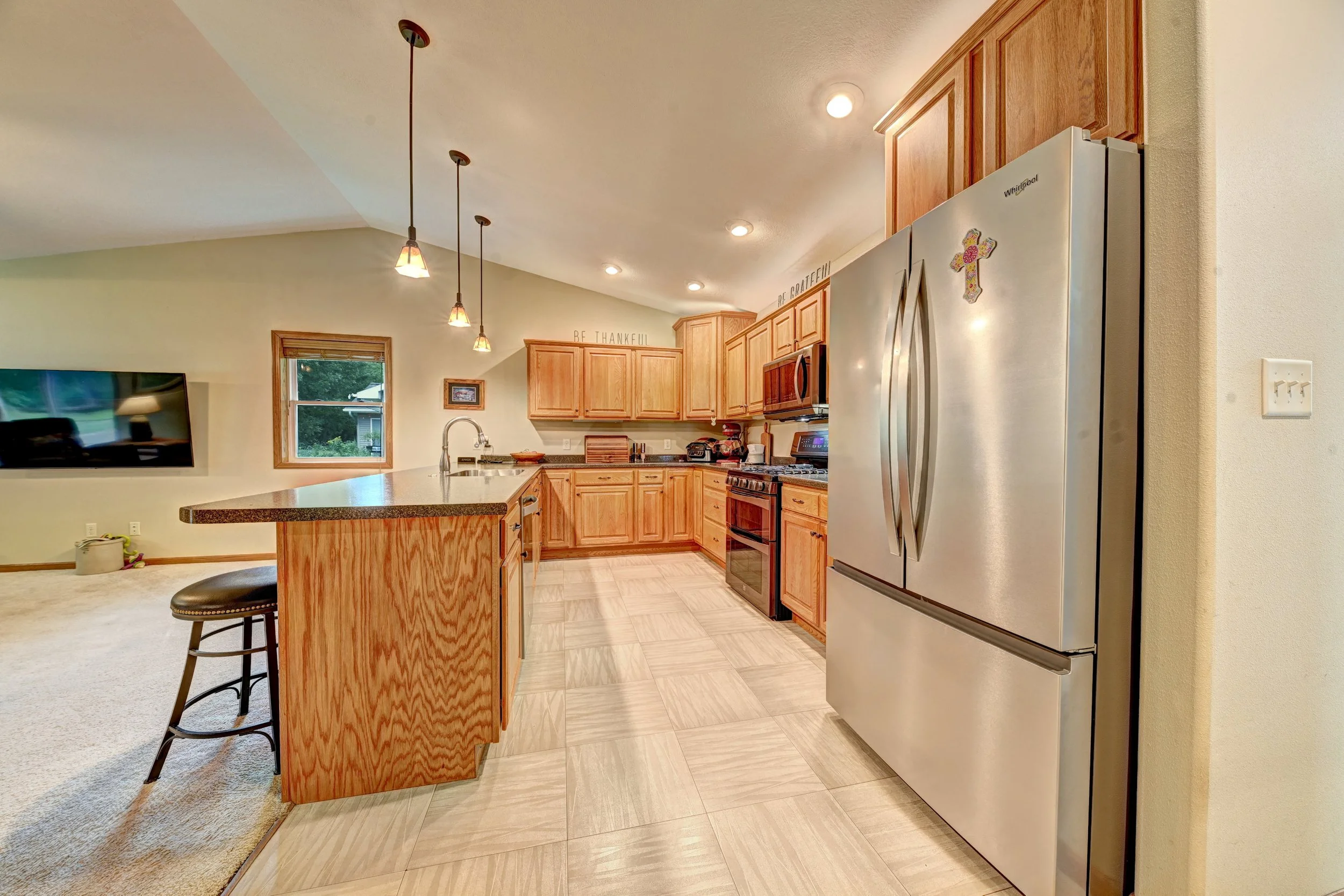Kitchen with wooden cabinets, stainless steel refrigerator with a cross magnet, tile floor, pendant and recessed lighting, window, and a breakfast bar with a chair.