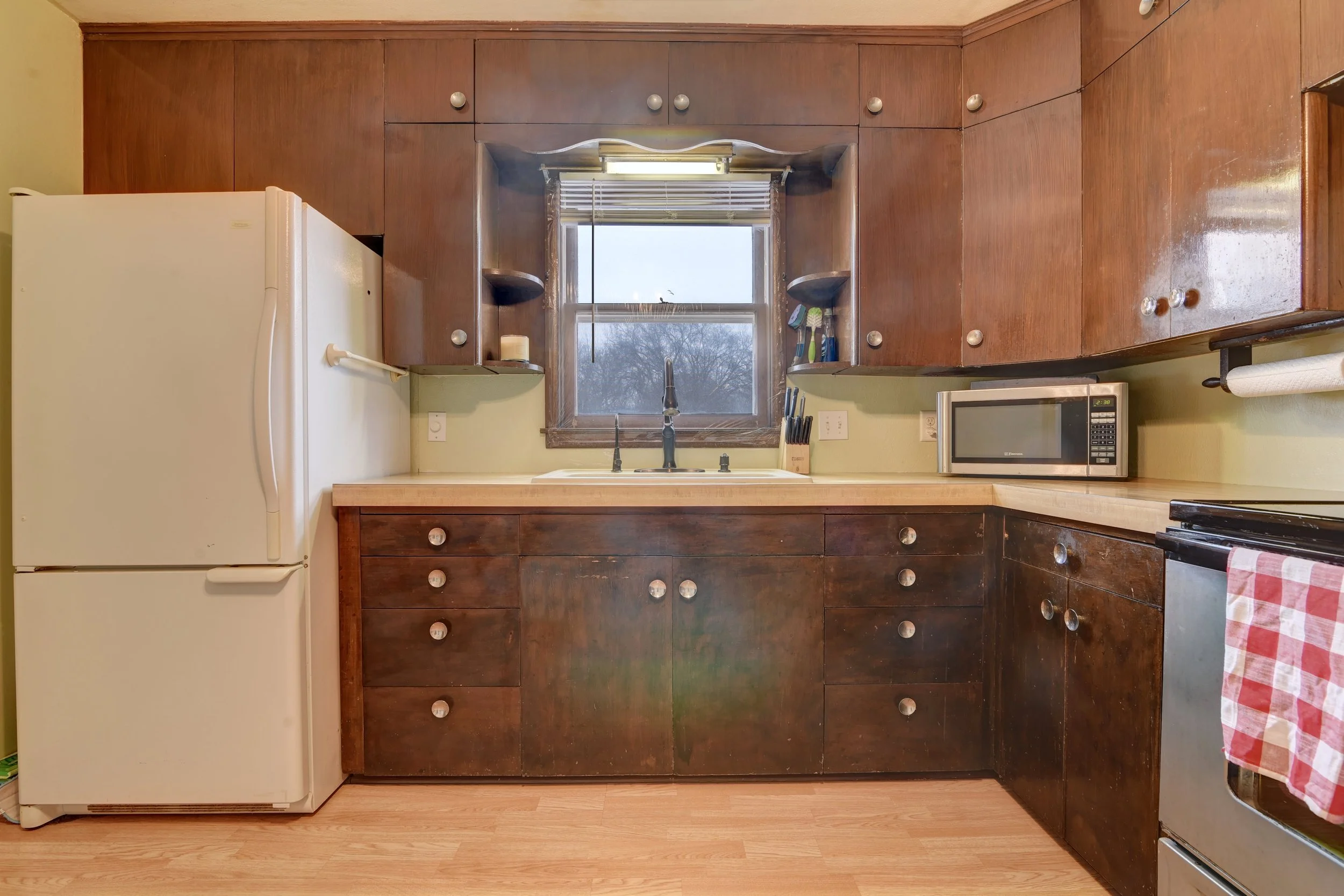 Kitchen with dark wooden cabinets, a white refrigerator, a window above the sink, a microwave on the countertop, and a stove with a red and white checkered towel hanging.