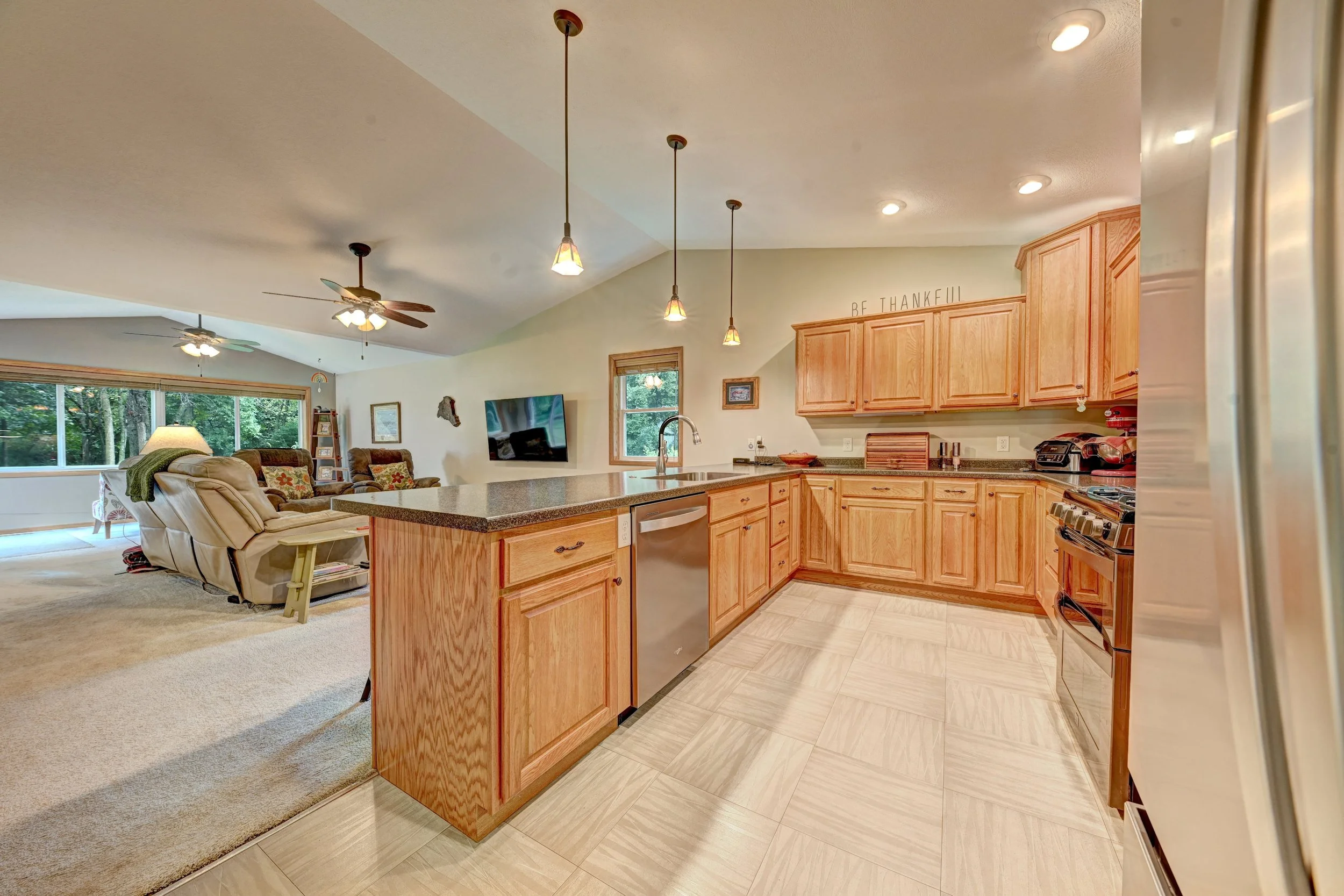 Open-concept kitchen with wooden cabinets, granite countertops, pendant lighting, and a view into the living room with large windows, ceiling fans, and furniture.