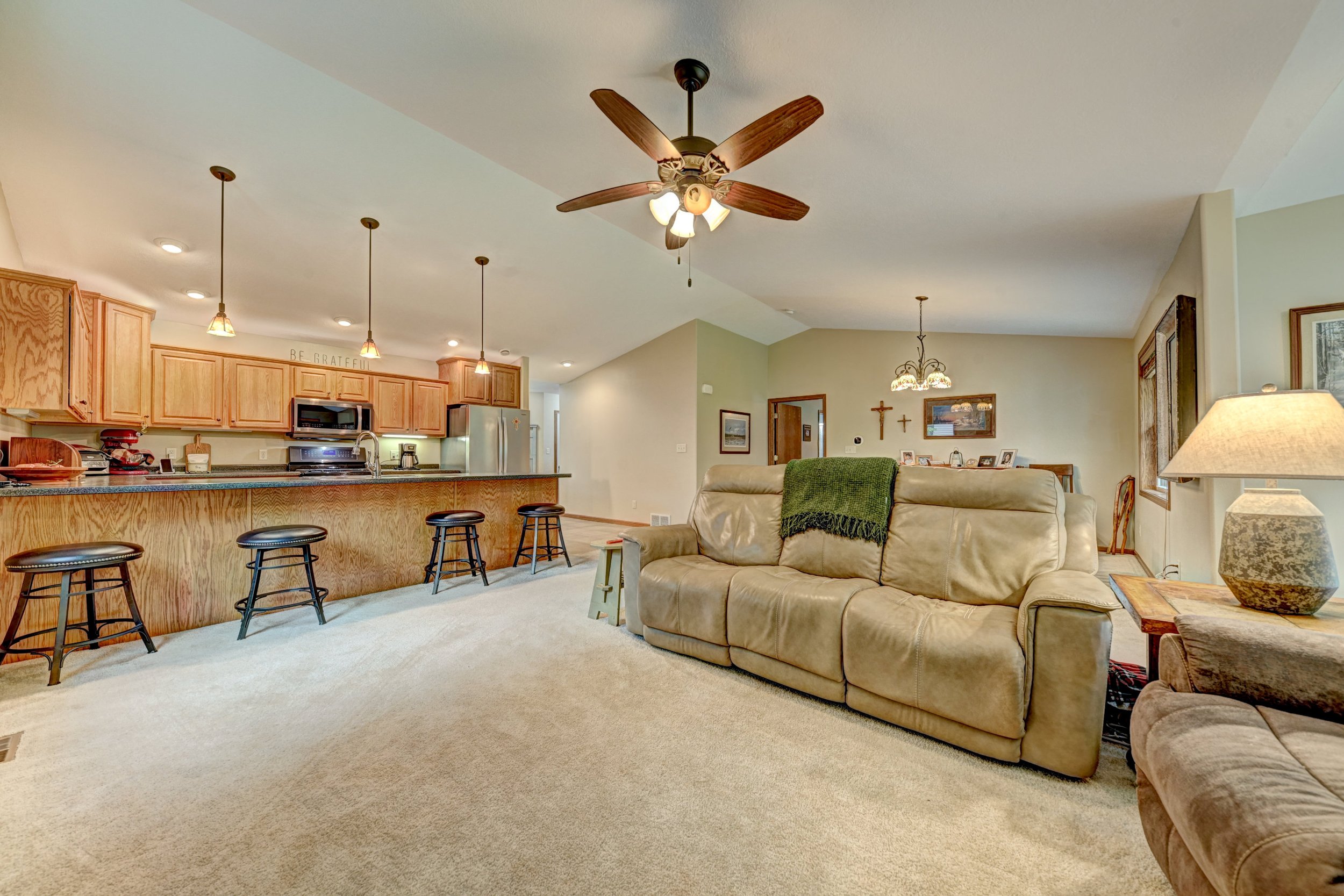 Open concept living room and kitchen with wooden cabinets, bar stools, ceiling fan, and various decorations.