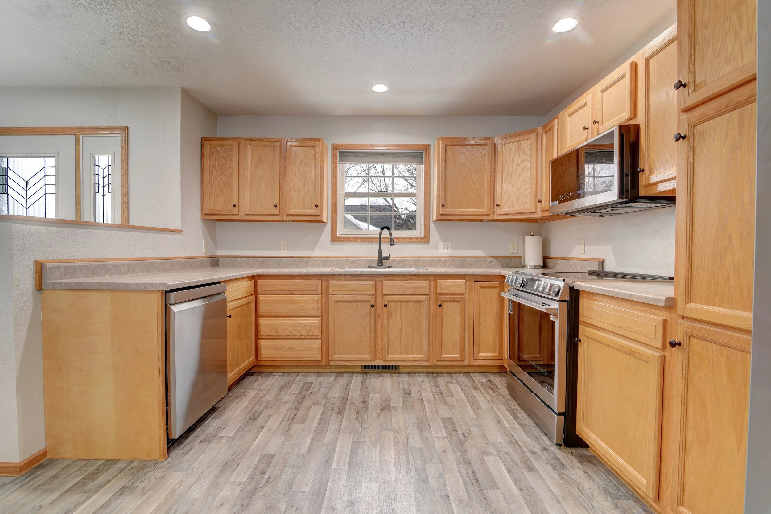 Empty kitchen with wooden cabinets, stainless steel appliances, a window above the sink, and laminate flooring.
