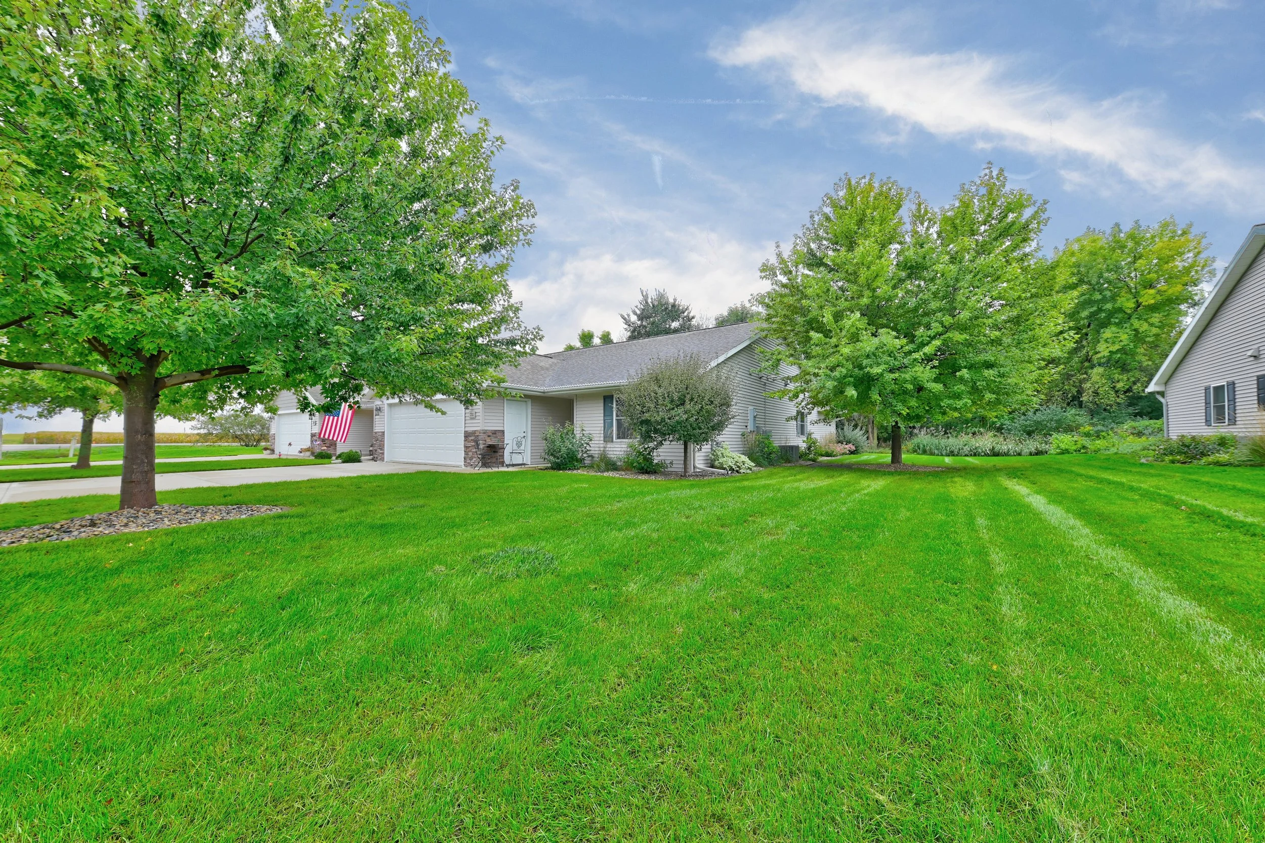 A suburban yard with green grass, large leafy trees, and a house with a white garage door. The house has beige siding, and there is an American flag hanging near the garage. The driveway is concrete, and the sky is partly cloudy with some blue visibl