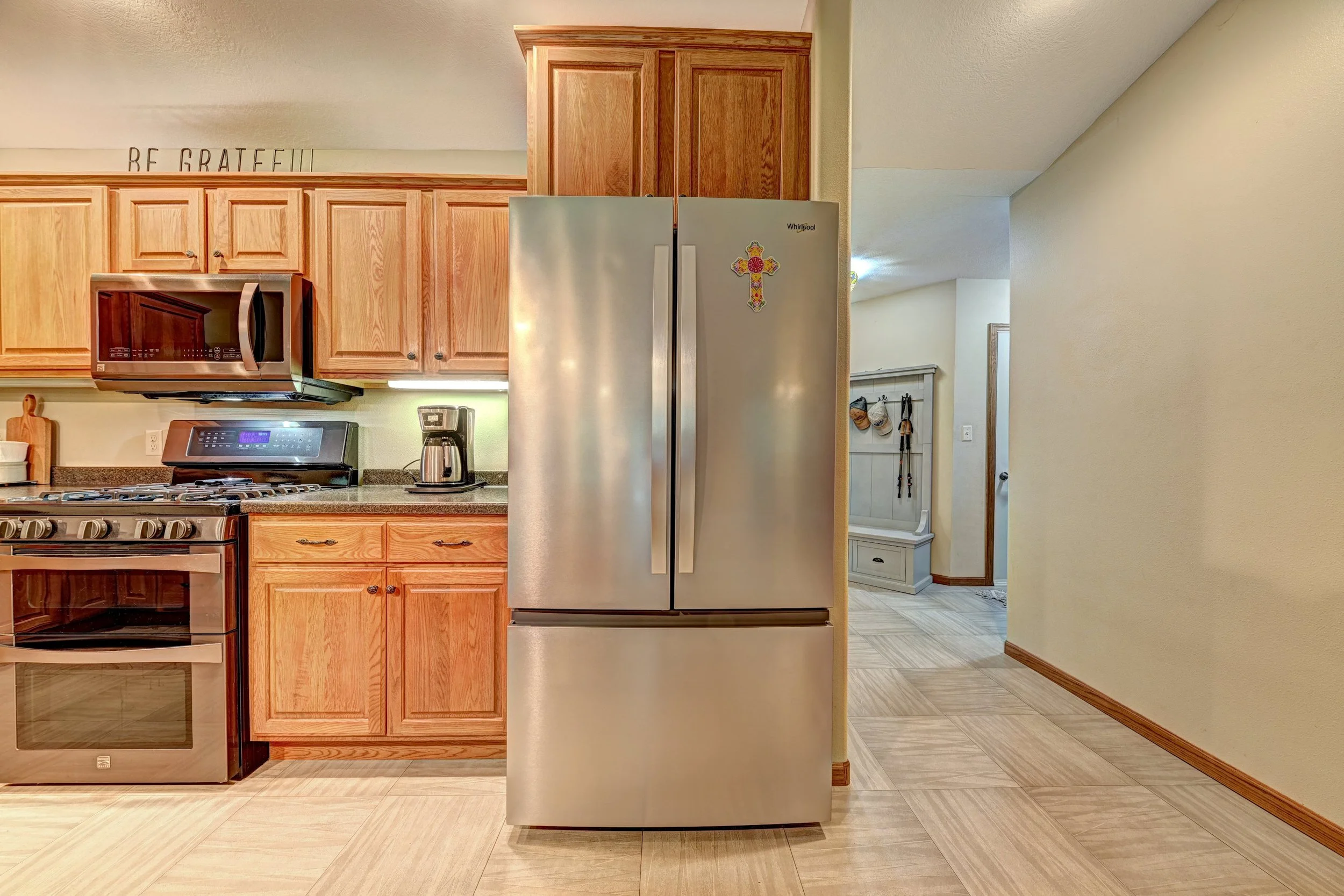Kitchen with wooden cabinets, stainless steel refrigerator with a cross magnet, microwave, stove, coffee maker, and a hallway with coat hooks.