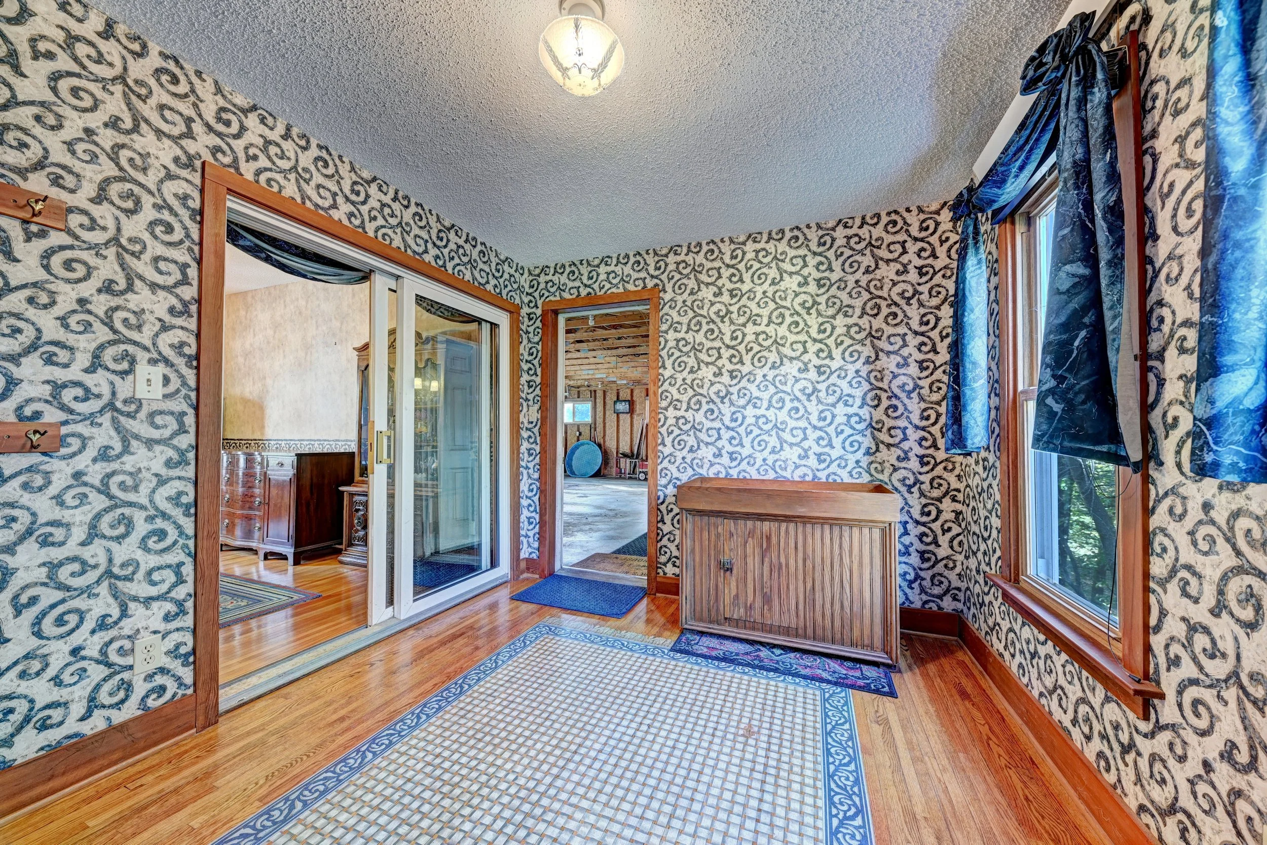 Interior view of a room with patterned wallpaper, hardwood floors, and a wooden coat rack. There are windows with curtains and a doorway leading to another room with wooden walls and a TV.
